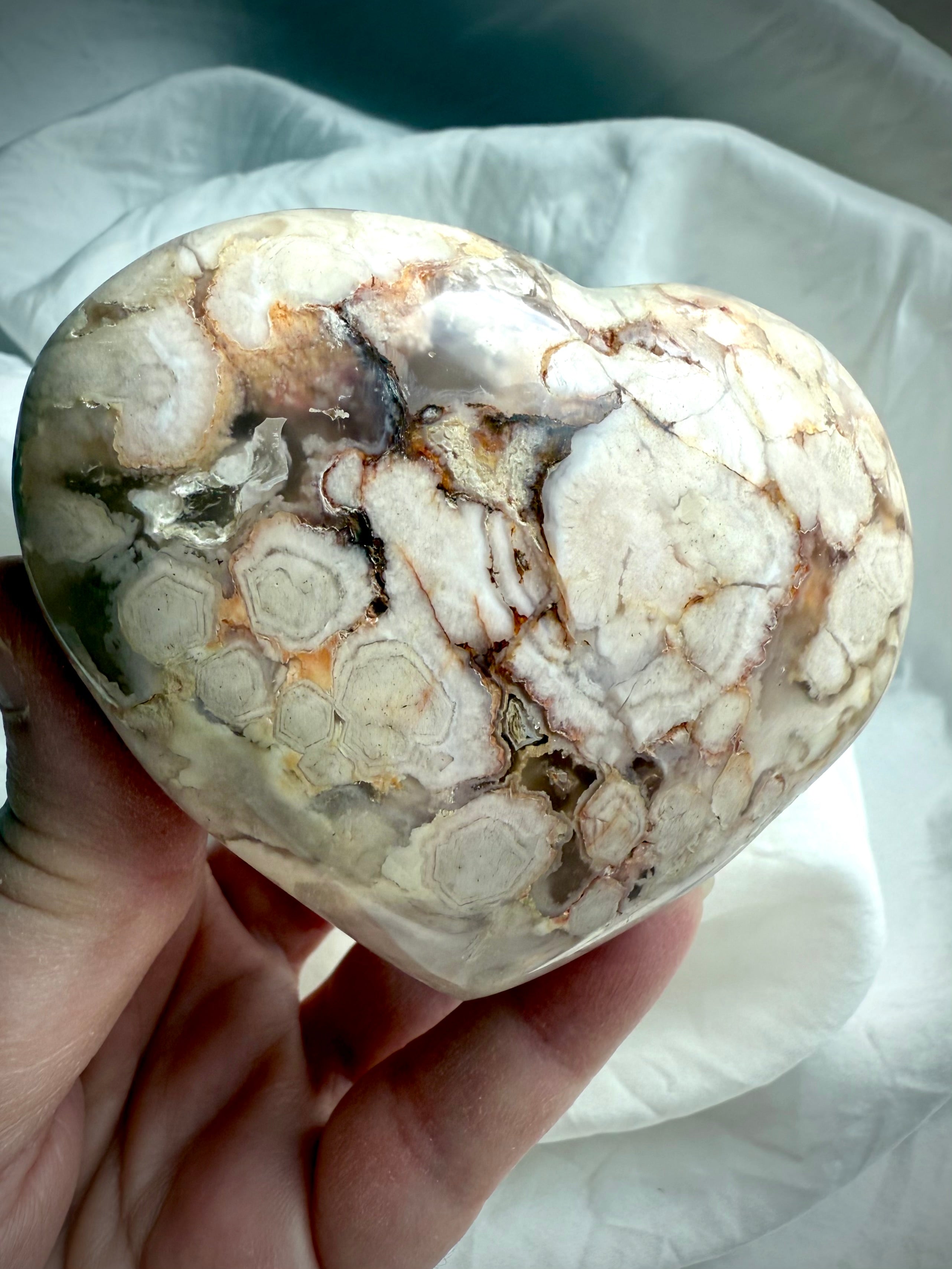 Hand holding a Flower Agate heart at an angle, capturing its blooming patterns and soft pastel hues — gentle crystal for emotional balance, resilience, and renewal.