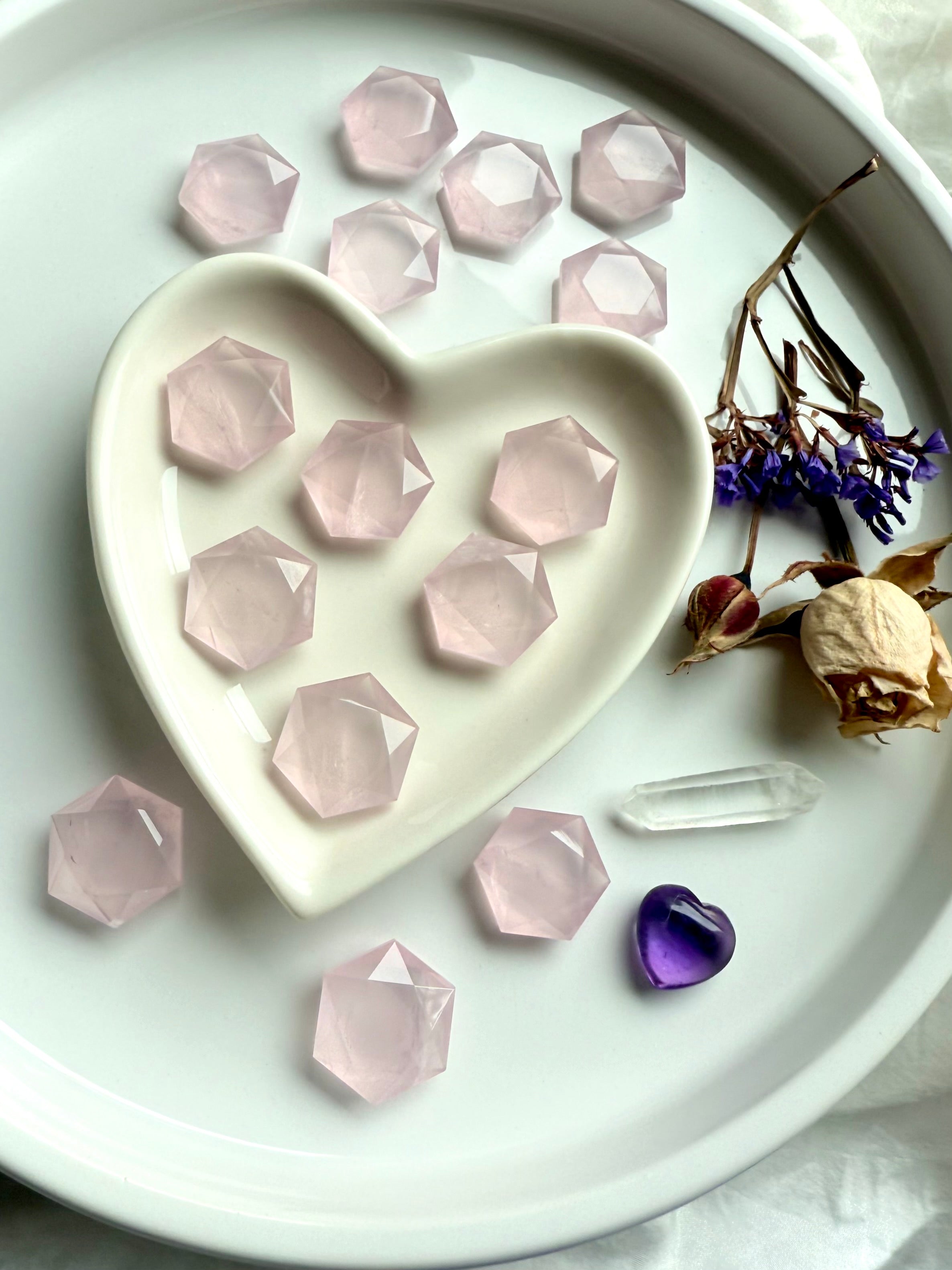 Mini rose quartz hexagon crystals displayed in a white heart bowl with more scattered on a plate, styled with roses, clear quartz point and amethyst heart