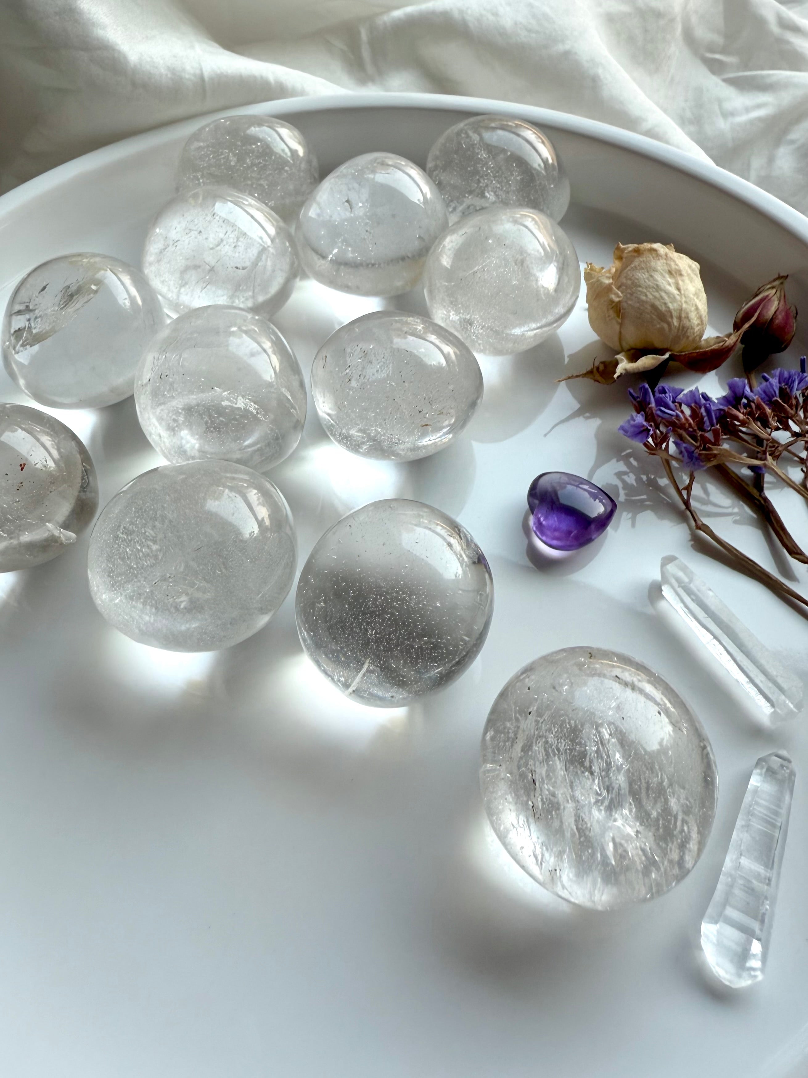 Angled view of Clear Quartz tumbled stones on a white plate with quartz points, amethyst heart, and flowers — Crown Chakra crystals for clarity, focus, and healing energy