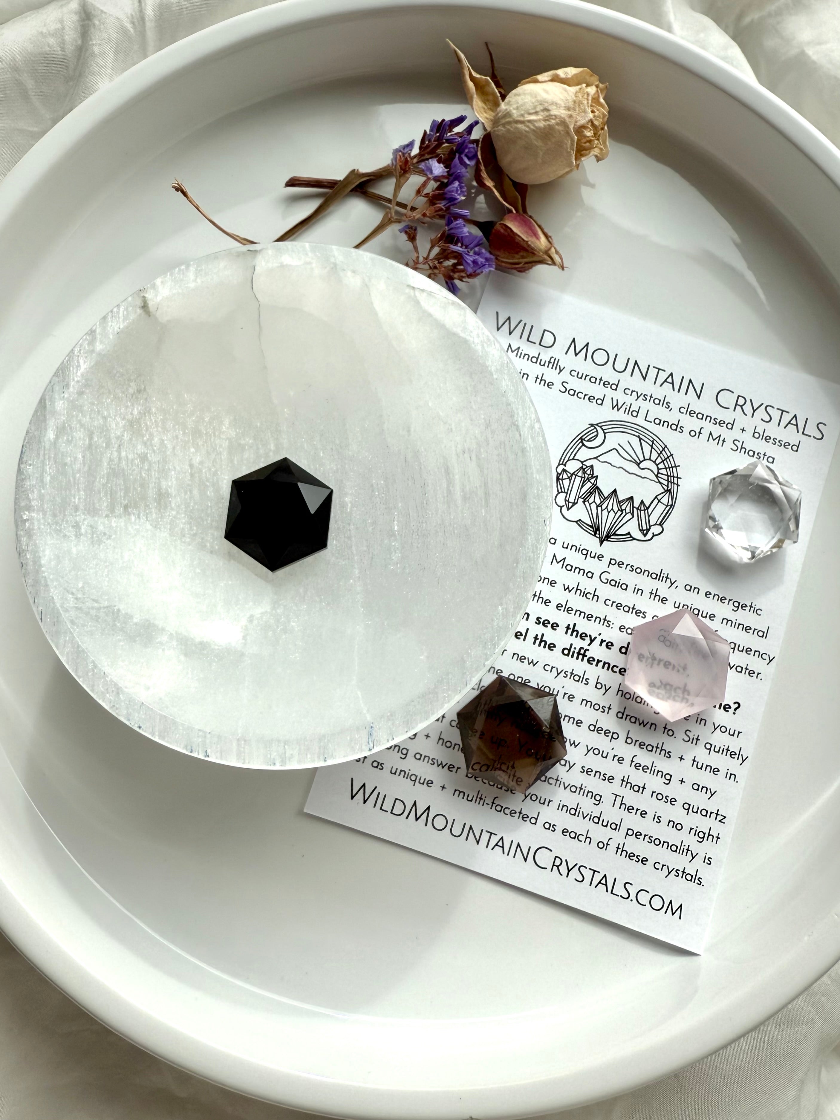 Mini faceted black obsidian hexagon displayed on a selenite disc with rose, smoky, and clear quartz hexagons, dried flowers, and Wild Mountain Crystals info card