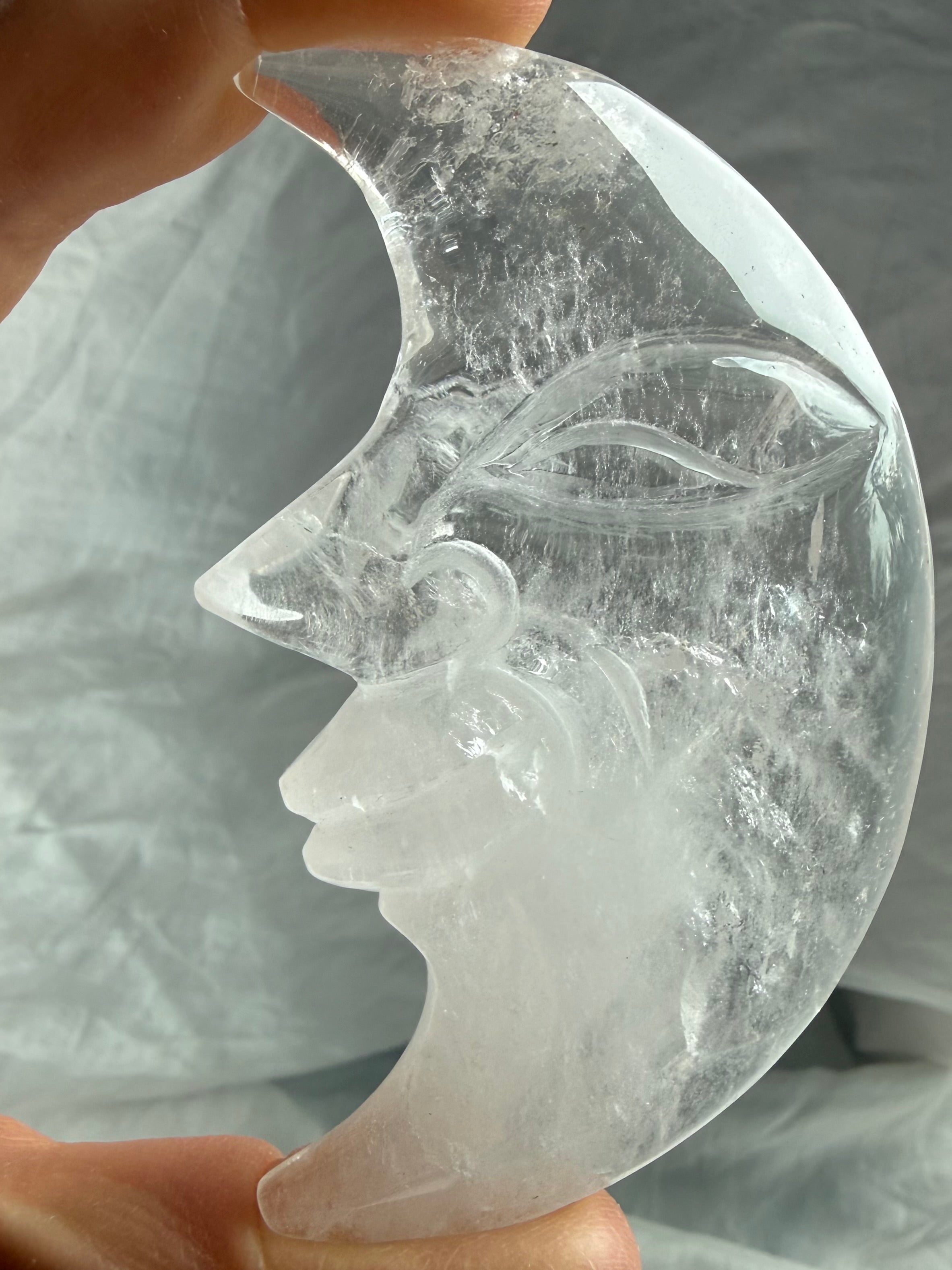Clear Quartz Moon Face carving held in natural light, revealing exceptional clarity, smooth glossy polish, and celestial design