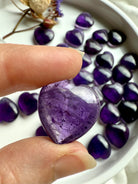 Amethyst heart held between fingertips showing its luminous purple hue — a crown chakra healing stone for intuition, clarity, and spiritual protection.