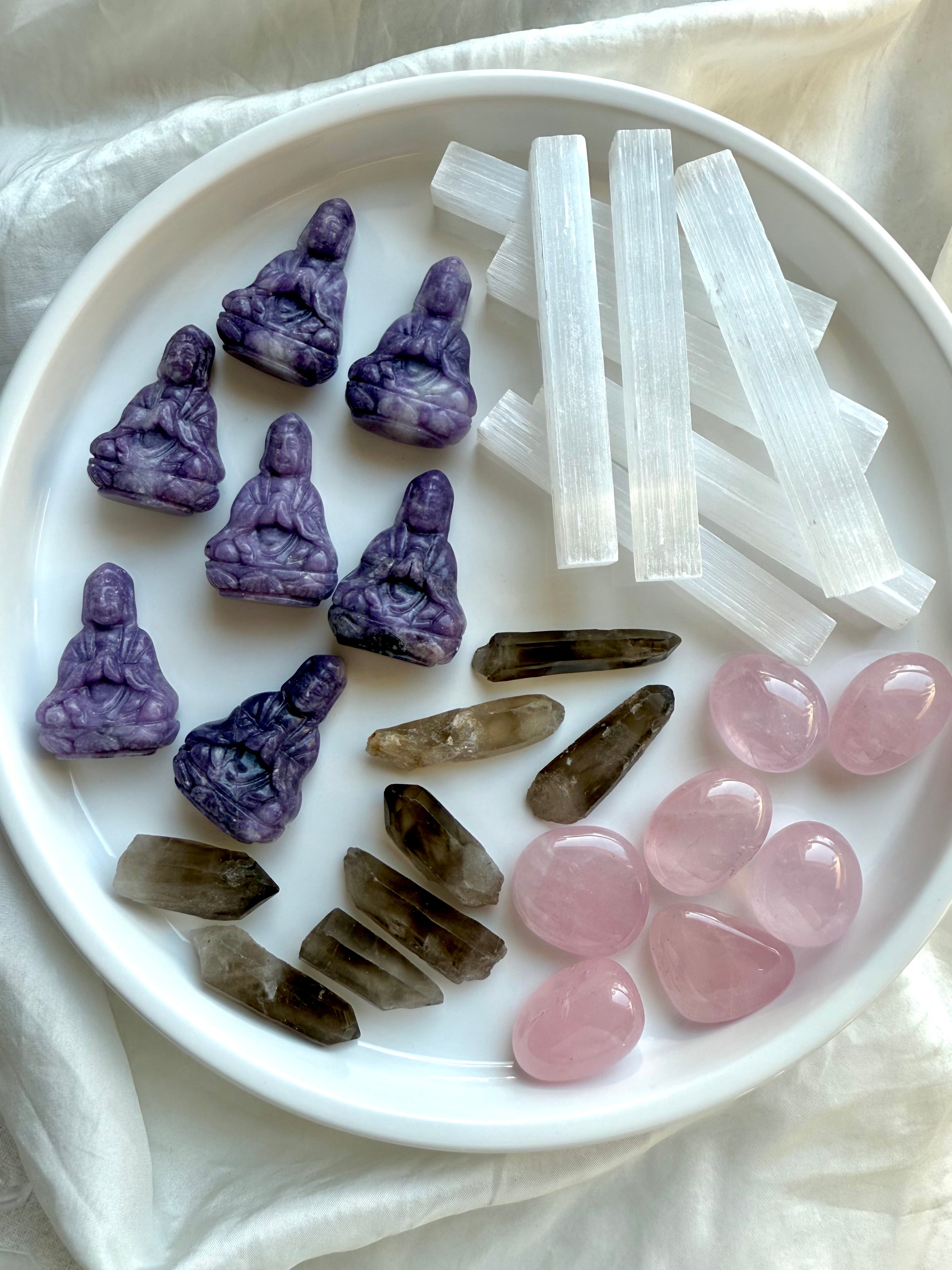Multiple sets of travel altar components: lepidolite Quan Yin figurines, raw smoky quartz, rose quartz stones, and selenite sticks arranged in white tray