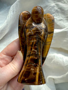 Hand holding Tiger’s Eye angel figurine with strong contrast in golden brown hues, emphasizing its guardian shape