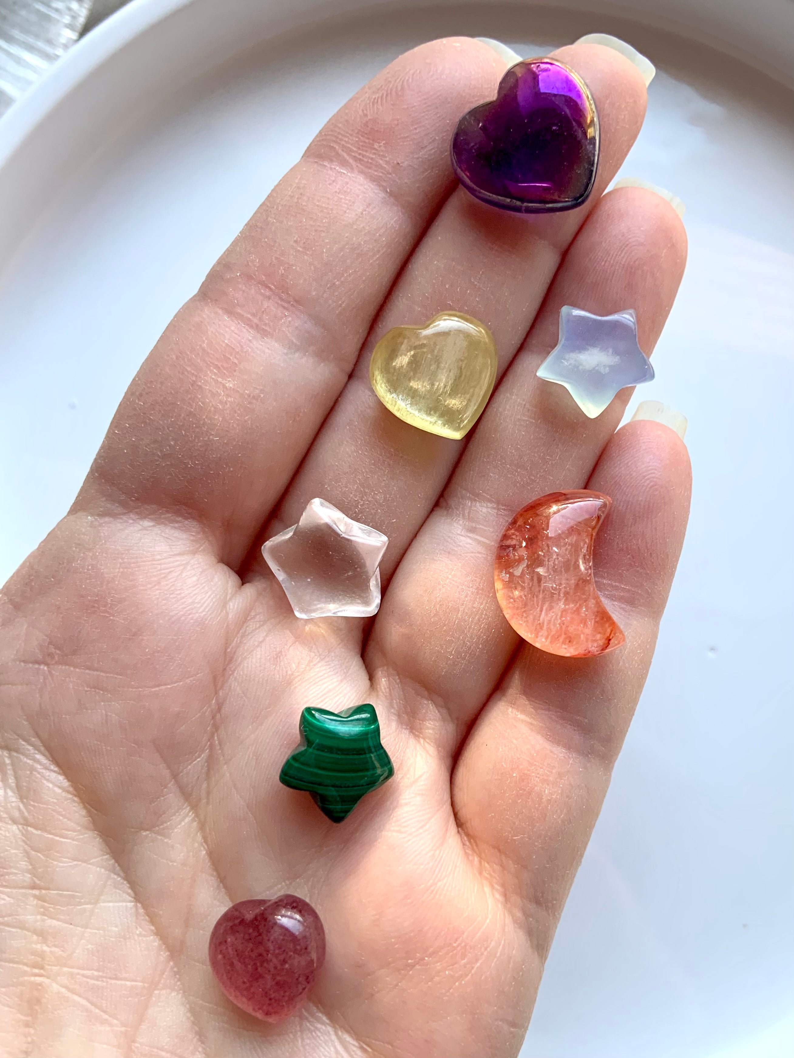 Hand holding 7 carved chakra crystals in heart, star & moon shapes in rainbow colors for each chakra