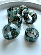 polished moss agate wide band rings grouped together on marble surface with translucent depth and organic natural patterns