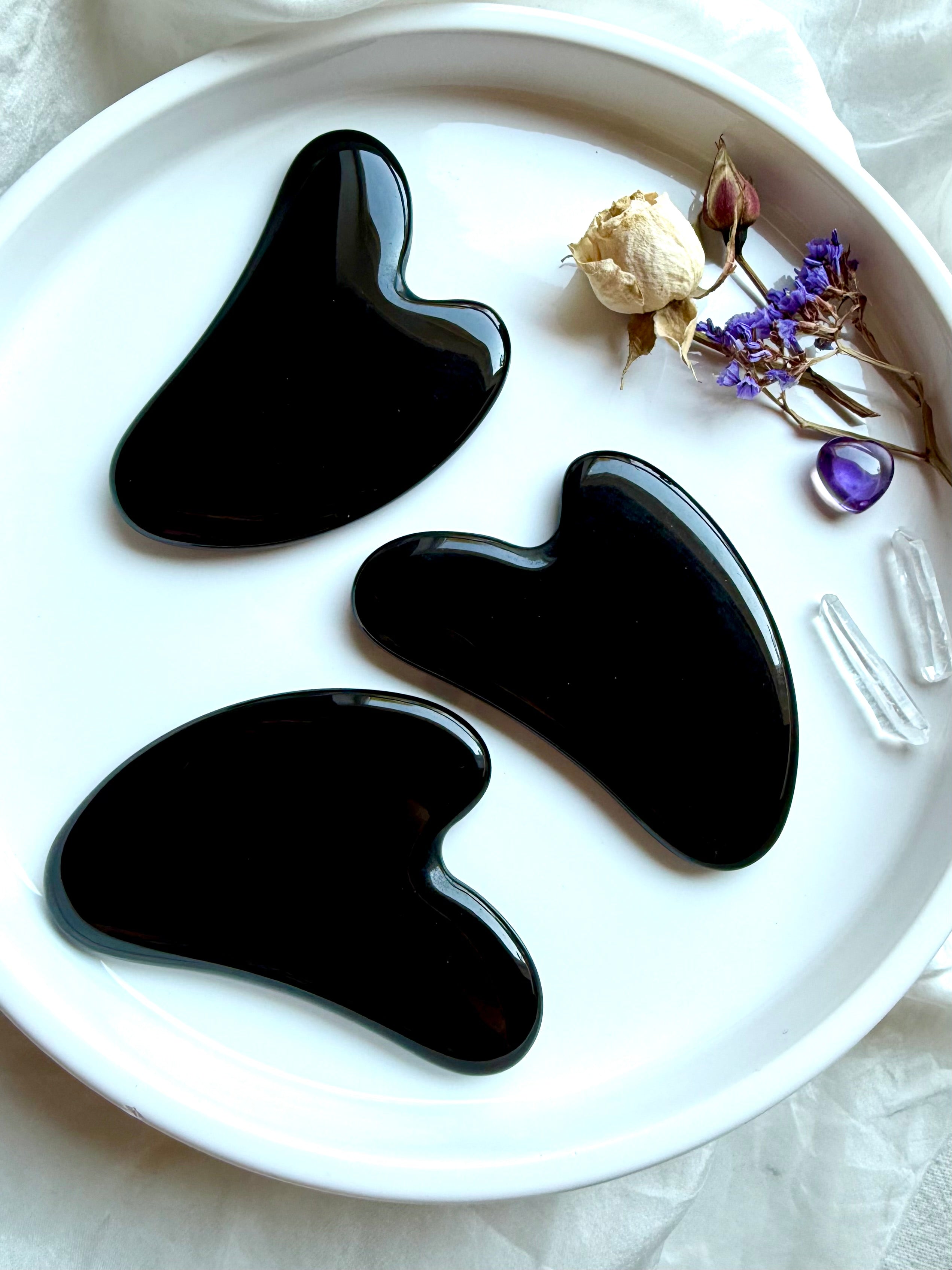 Three Black Obsidian crystal gua sha facial massage tools on white plate with dried flowers and amethyst accent