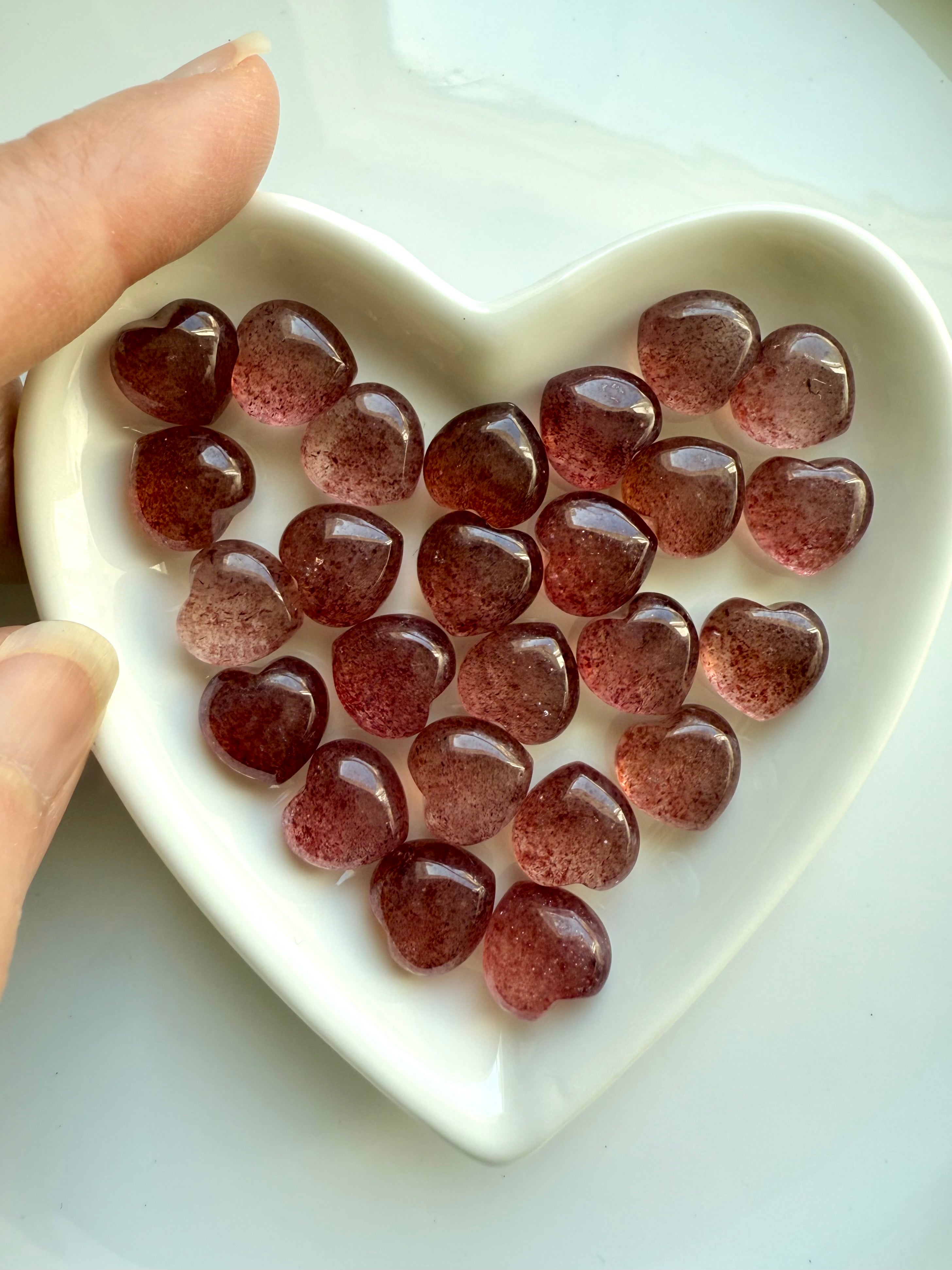 Mini Strawberry Quartz hearts in a white ceramic heart bowl with a hand reaching in — sweet crystal gift for love and emotional healing.
