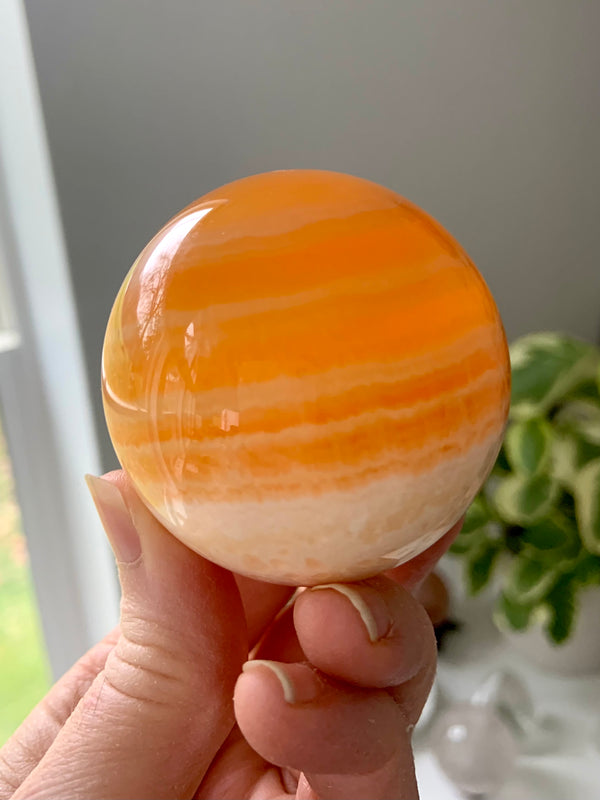 Translucent Orange Calcite Sphere with white banding, shown held on my hand in natural light