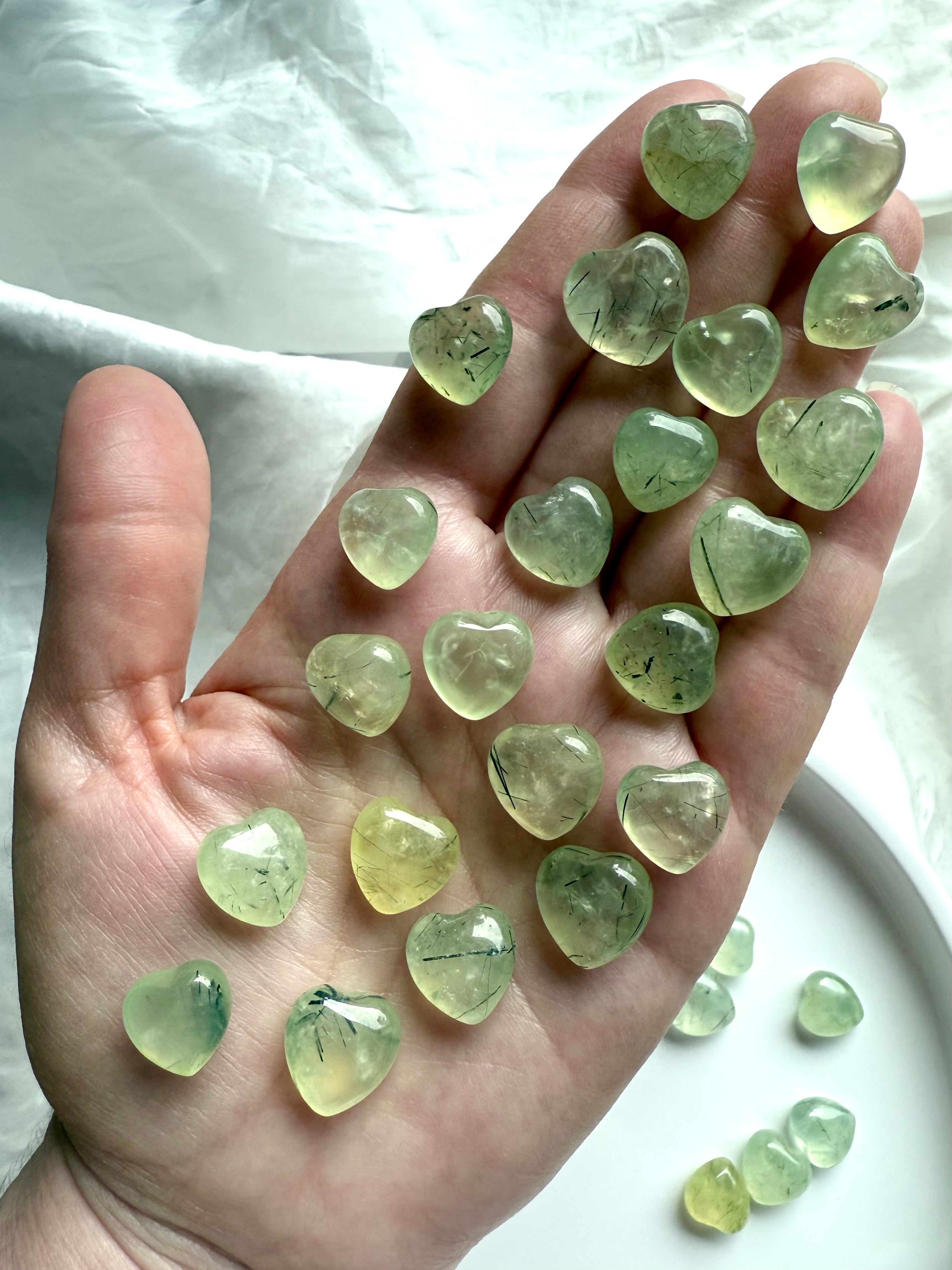 Hand holding multiple Prehnite crystal hearts with green translucence and needle-like epidote inclusions.