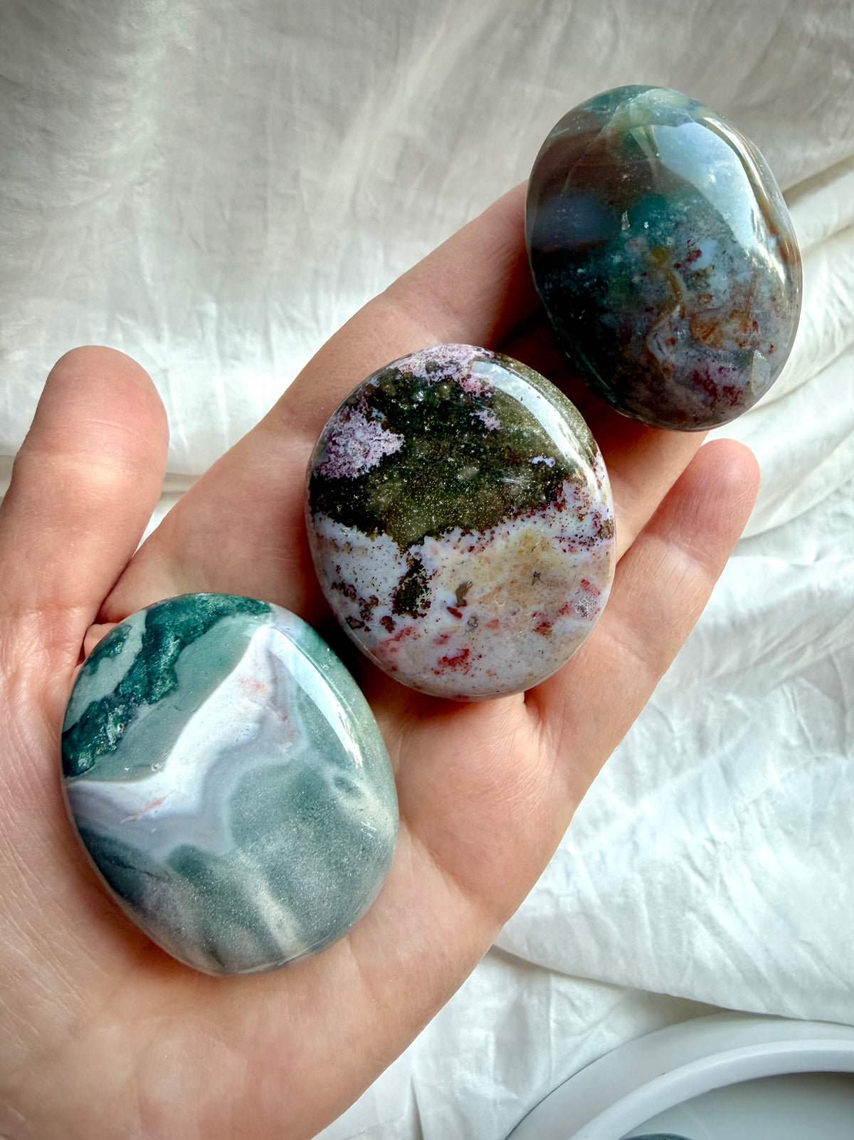 Three polished Ocean Jasper palm stones held in hand, showing green, gray, pink, and mauve Sea Jasper crystal patterns