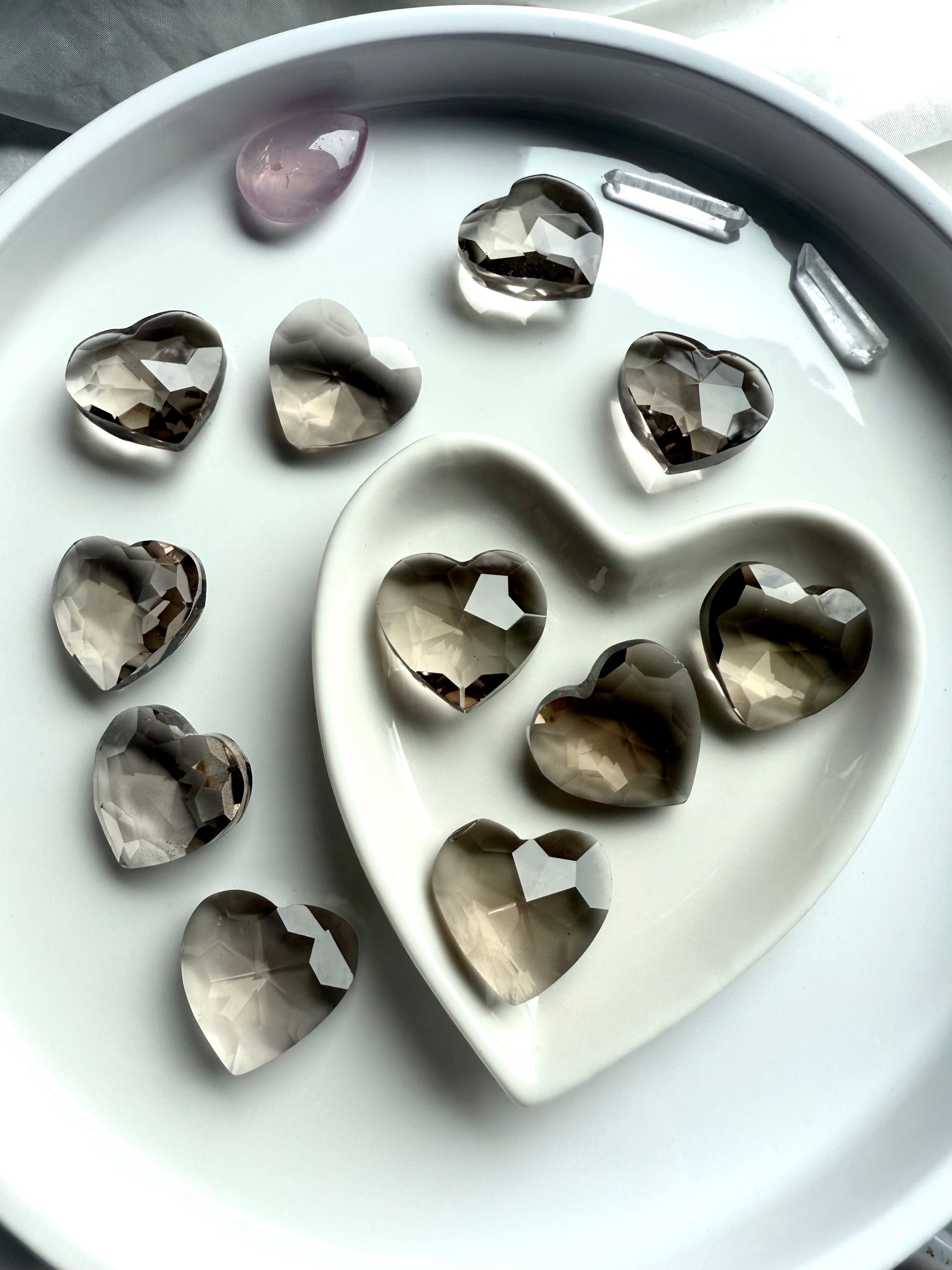 Faceted Smoky Quartz crystal hearts in a white ceramic heart bowl surrounded by more — spiritual gifts for grounding, stress relief, and emotional balance.