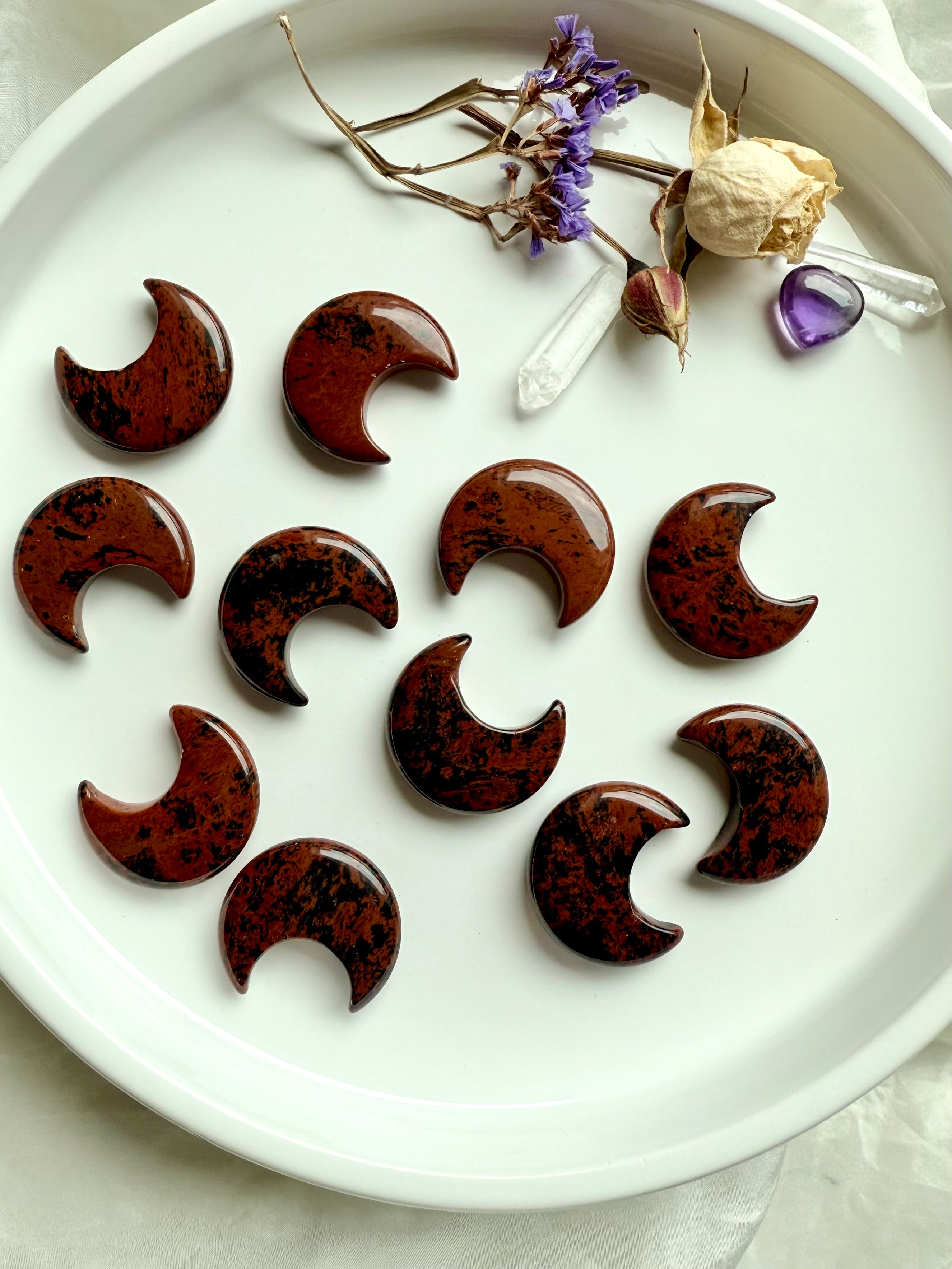 Long view of Mahogany Obsidian crescent moons displayed in rows on a white plate with flowers and crystals. Moon carvings for Root + Sacral Chakra healing.