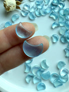 Closeup of two tiny Aquamarine crescent moon carvings resting on fingers, with a background of mini moons and dried flowers in soft lighting