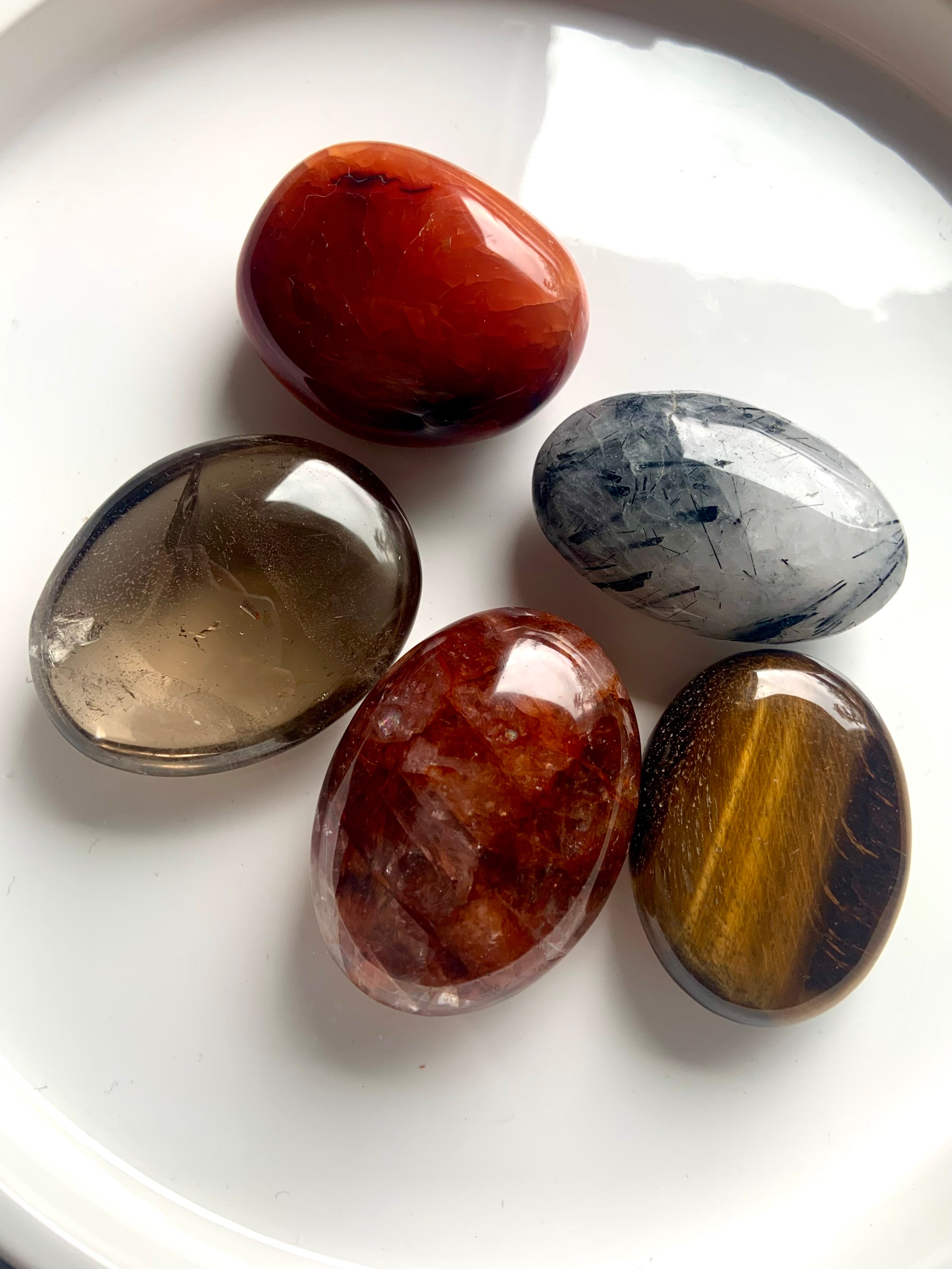 Closeup of five palm stones on white plate — includes Smokey Quartz, Tigers Eye, Carnelian, Fire Quartz &amp; Tourmaline in Quartz for chakra healing.