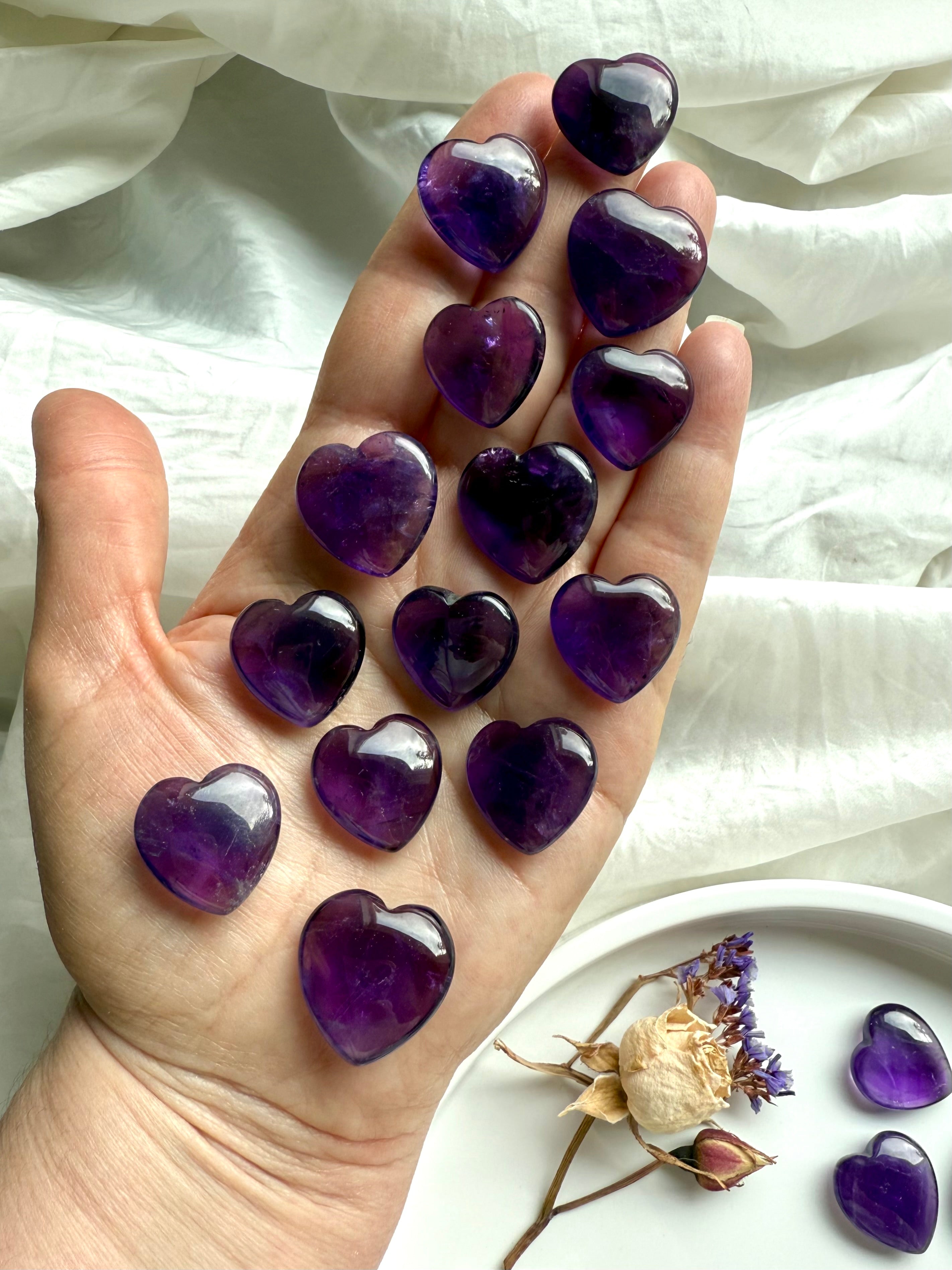 Amethyst crystal hearts in rich purple hues displayed in an open hand — polished crown chakra stones for intuition, clarity, and spiritual connection.