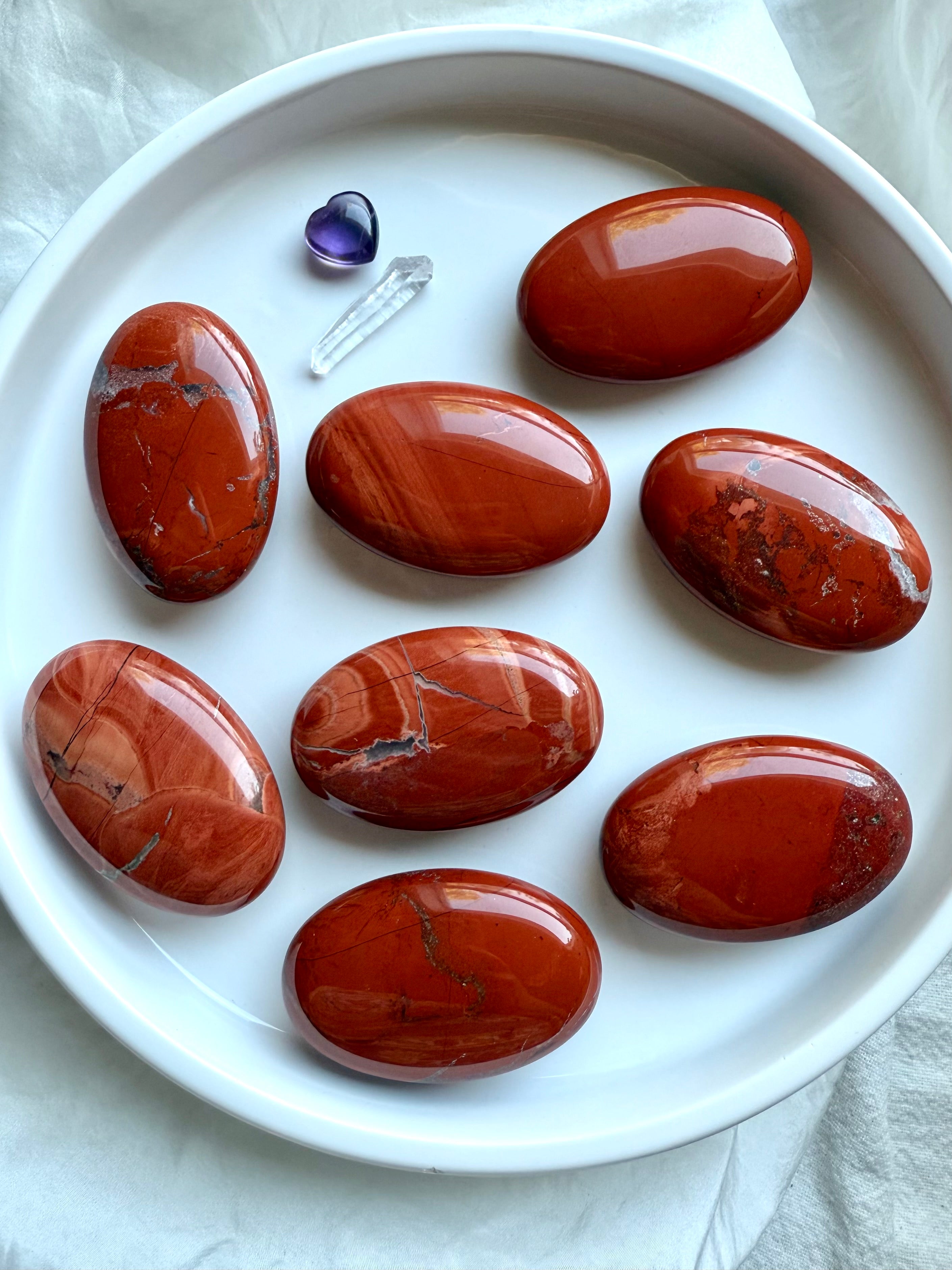 Flatlay of Red Jasper palm stones on white plate — smooth oval crystals with natural color variation.