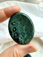 Kambaba Jasper worry stone held in hand with forest green, dusky teal, and black eye-like patterns for grounding and meditation
