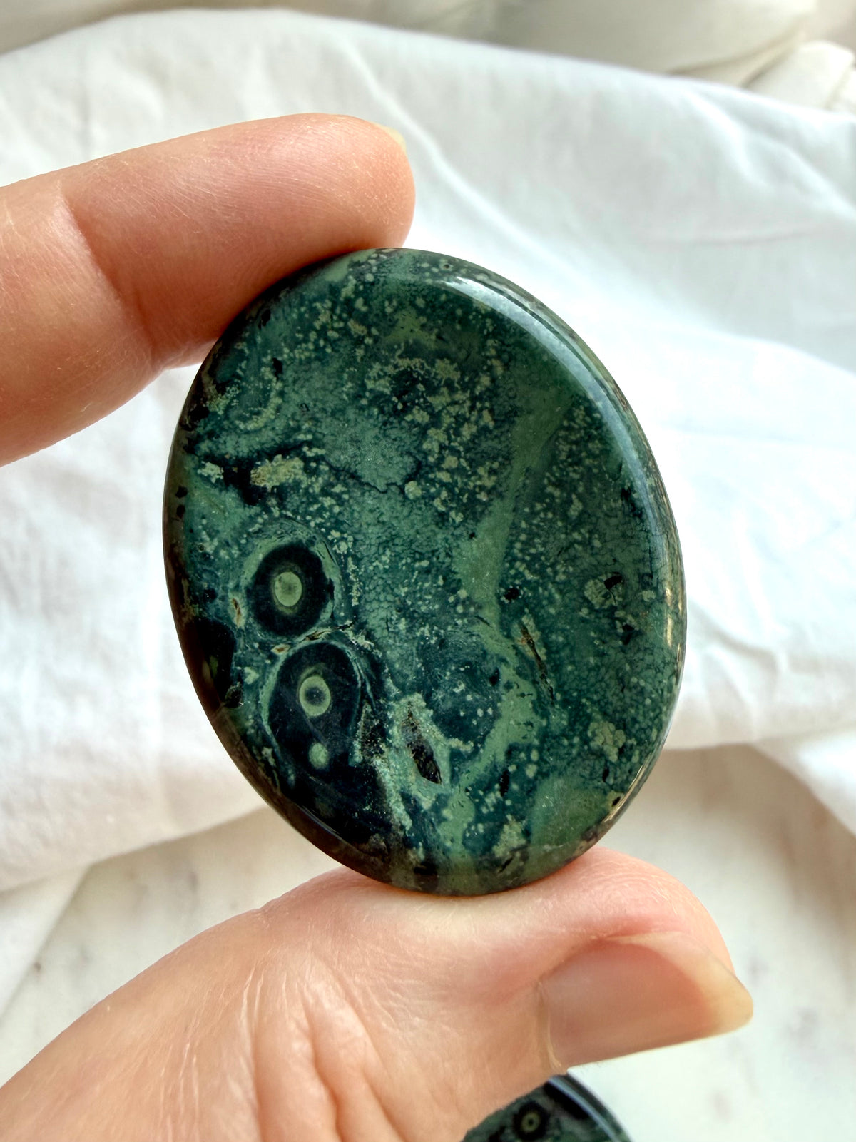 Kambaba Jasper worry stone held in hand with forest green, dusky teal, and black eye-like patterns for grounding and meditation