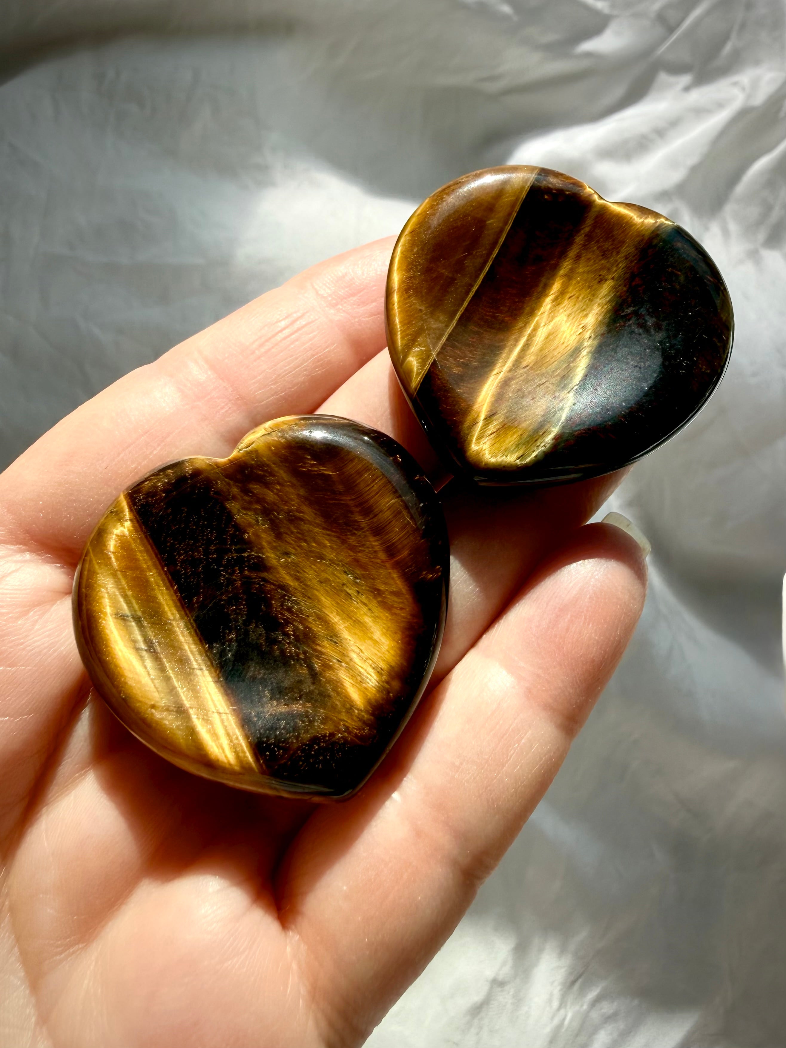Two polished Tiger’s Eye heart worry stones held in hand, featuring shifting golden light and natural striped patterns