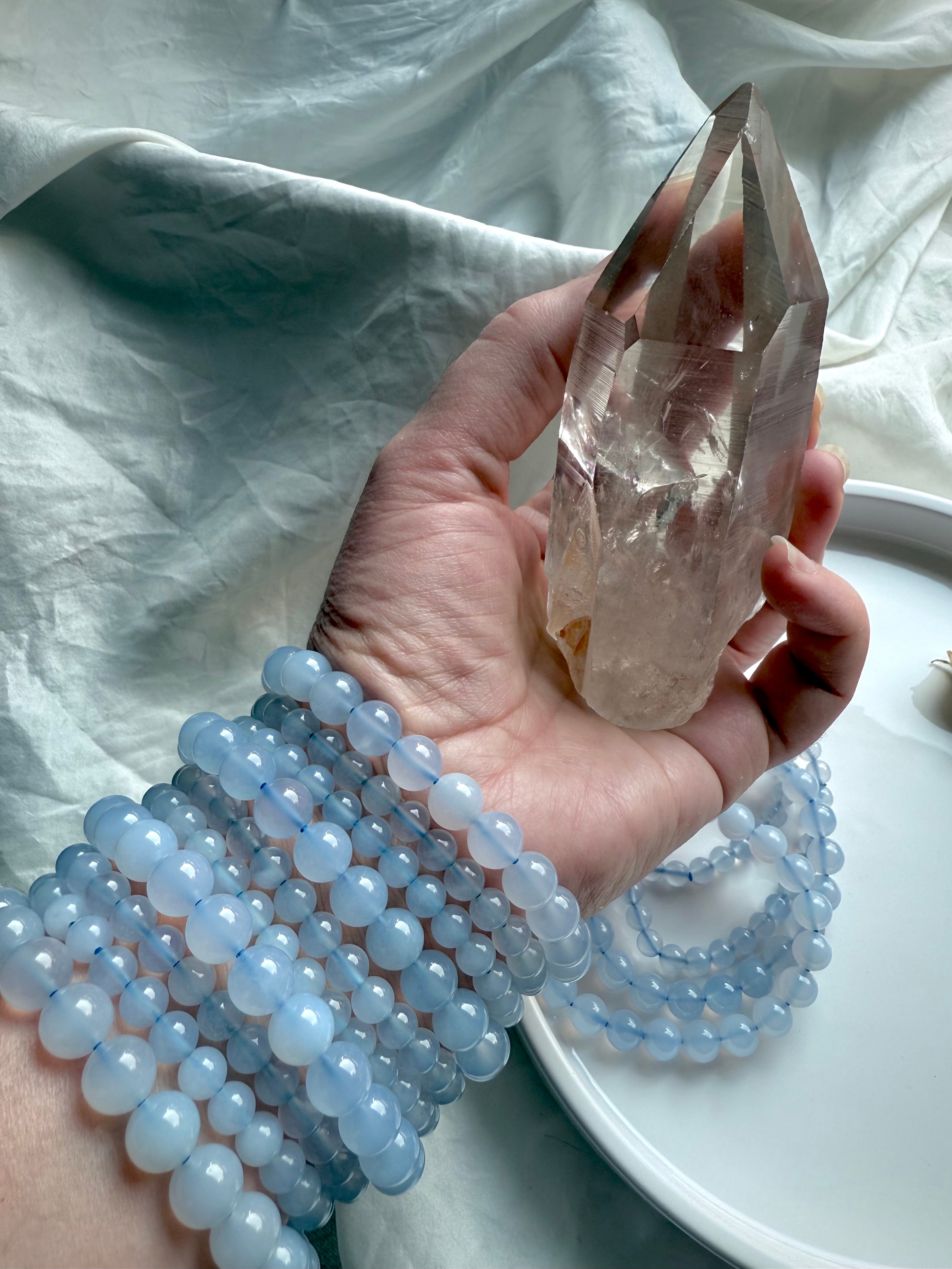Hand holding a smoky quartz point with a stack of Blue Chalcedony bracelets — throat chakra healing stones with gentle calming energy.