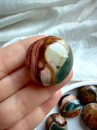 Mini Polychrome Jasper palm stone with deep green, cream, and rust red landscape patterns, held between fingers