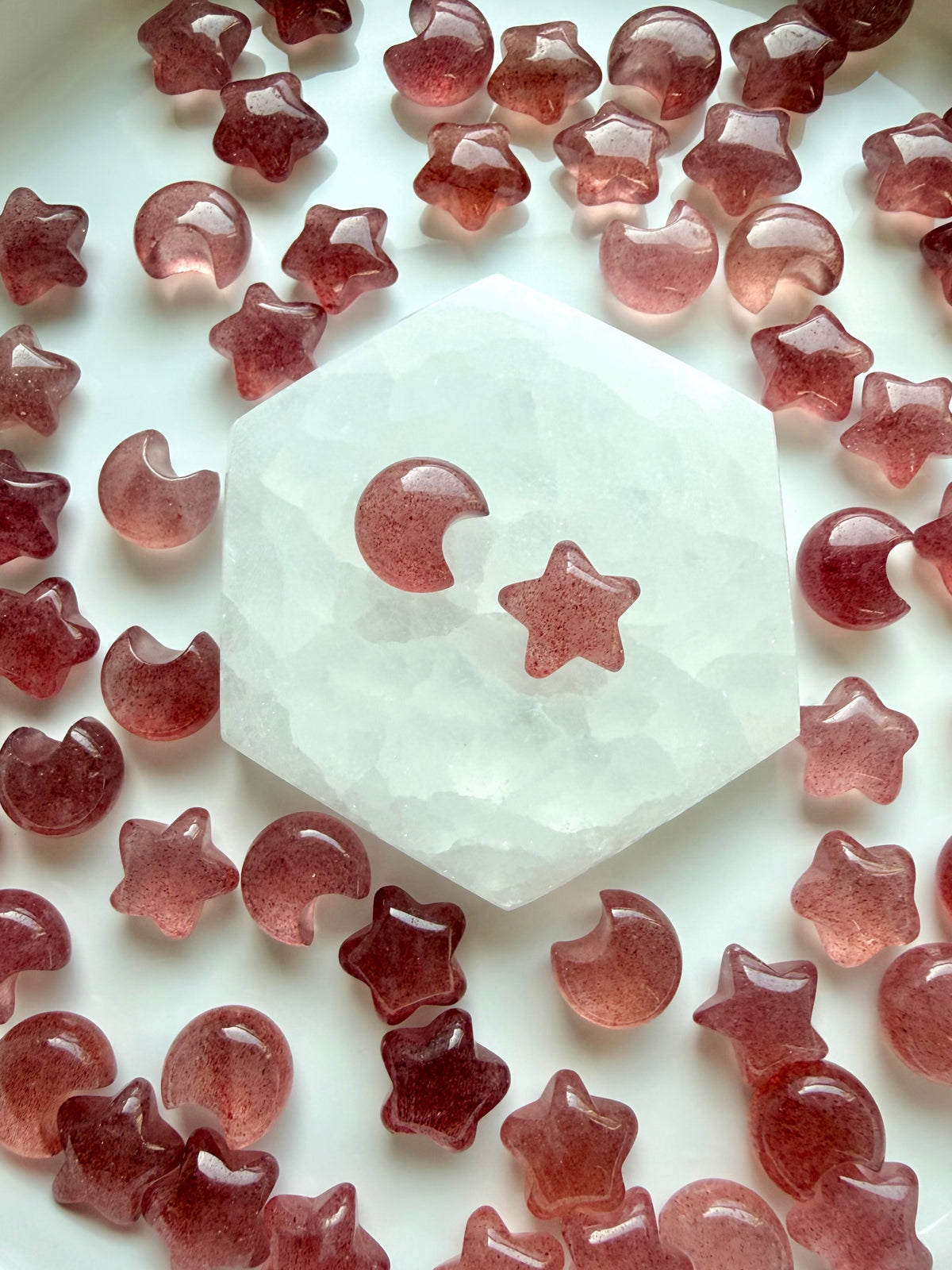 Strawberry Quartz mini moon and star carvings in translucent pink and deep berry red, displayed around a hexagonal Selenite charging plate
