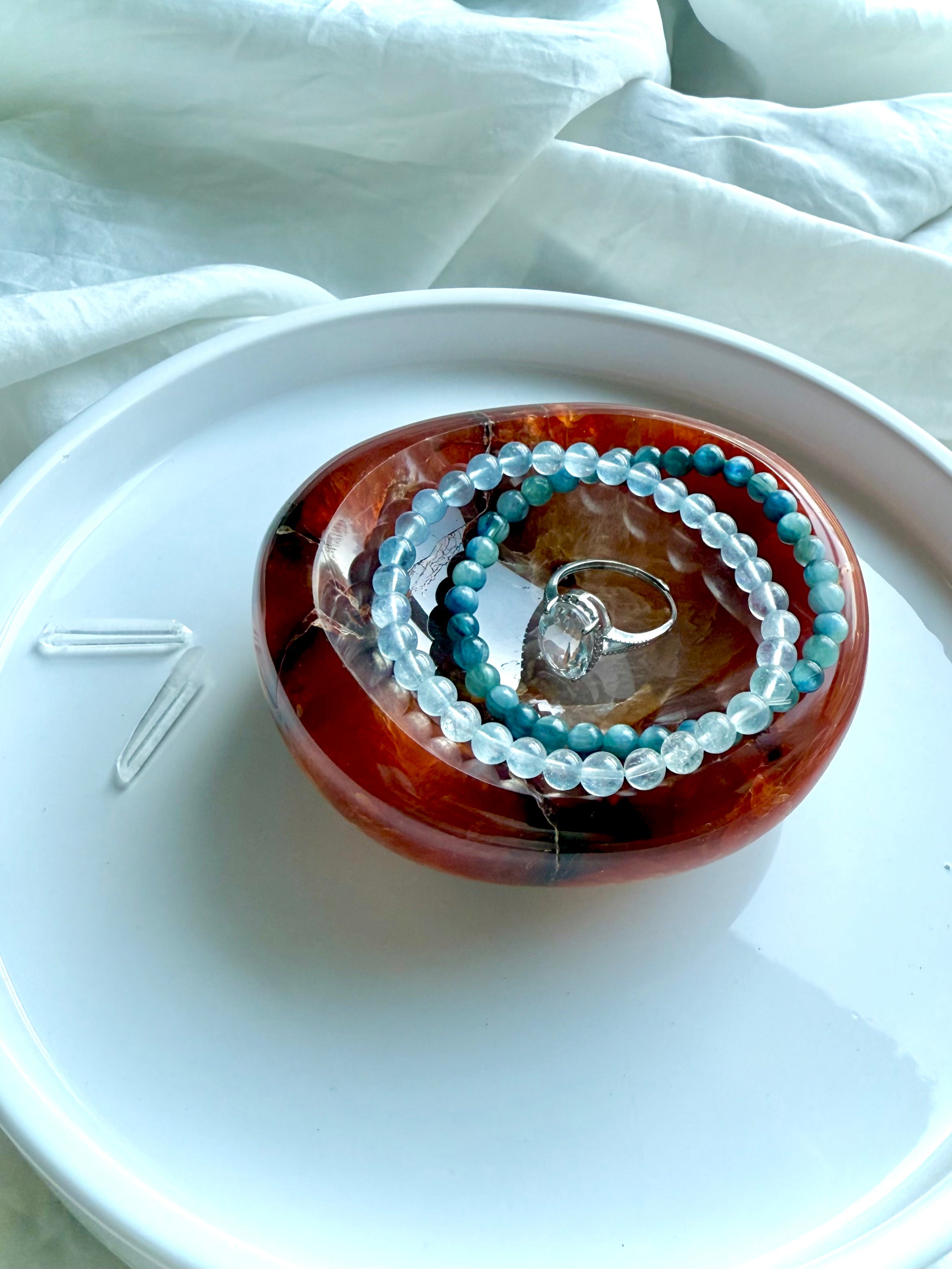 Carnelian crystal bowl used as a jewelry dish, holding aquamarine and kyanite bracelets with a silver ring — energizing gemstone decor for altar or sacred space