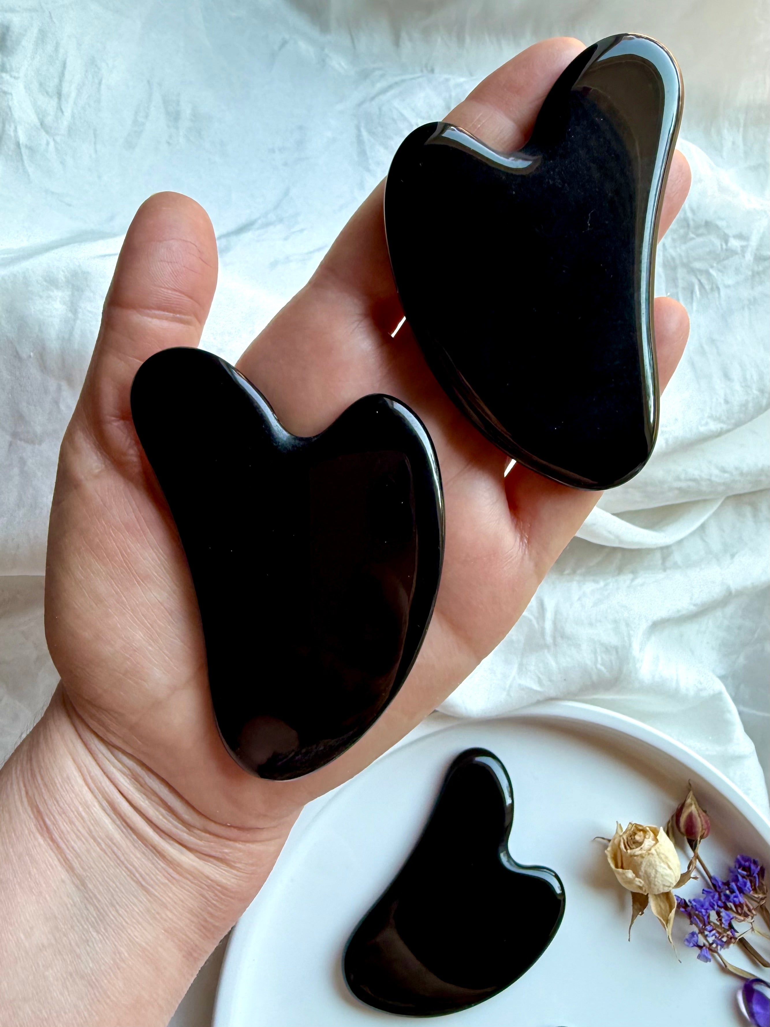 Hand holding two Black Obsidian crystal gua sha facial massage tools with curved contour shape, extra gua sha on plate below