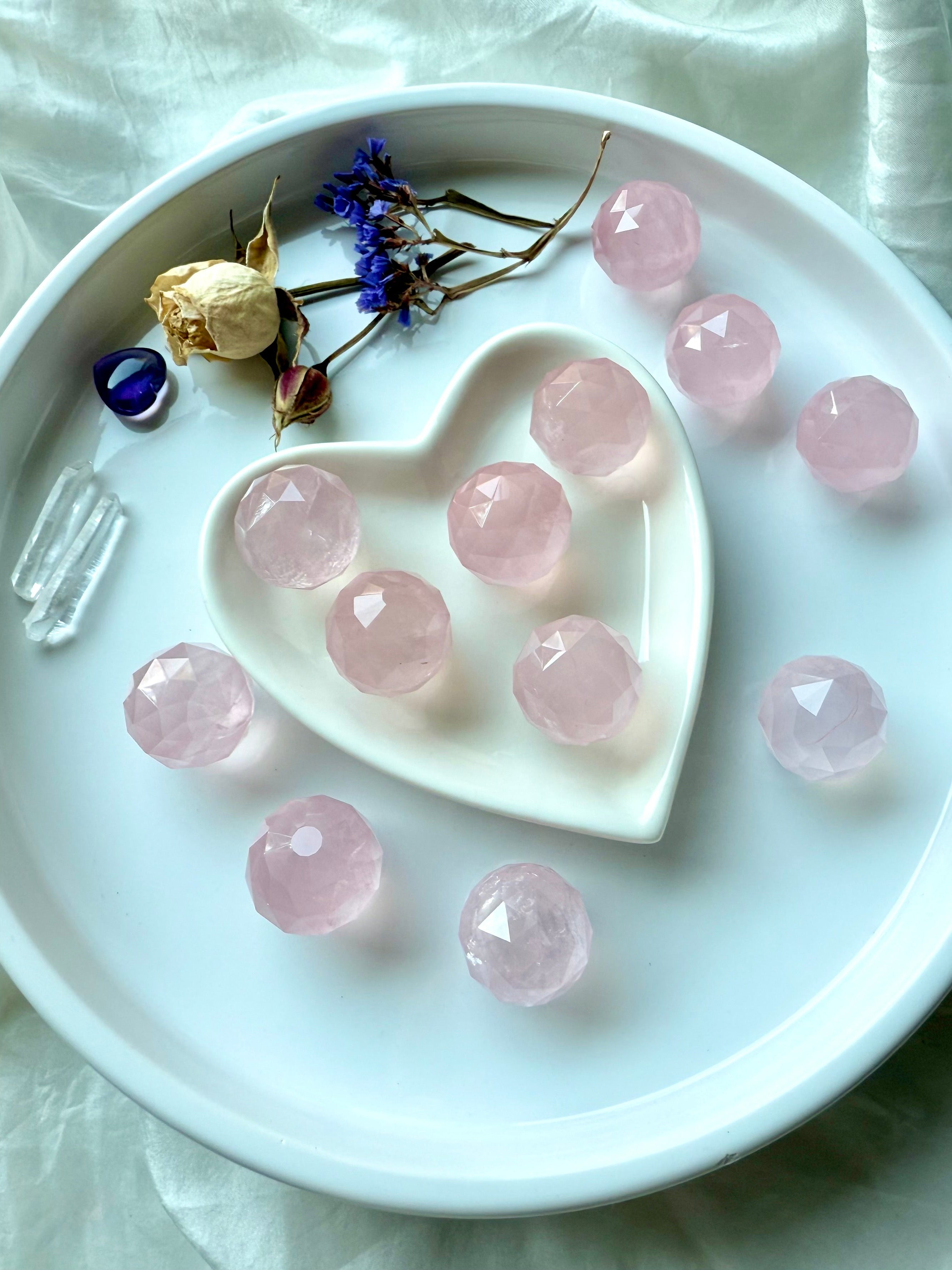 Faceted rose quartz mini spheres in a white heart-shaped dish with dried flowers and crystals. Soothing pink crystals for peace.