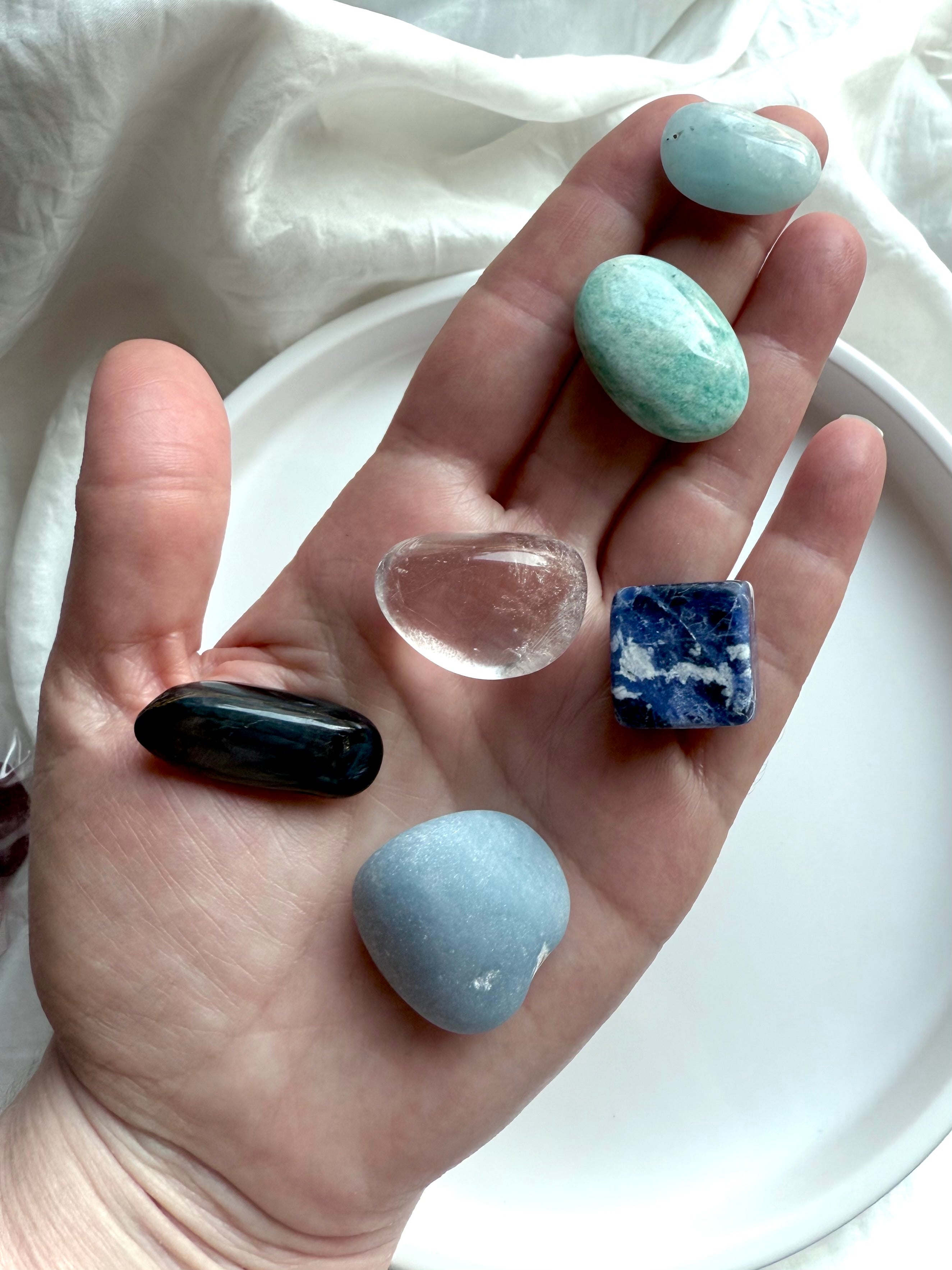 Hand holding six throat chakra crystals — Angelite, Sodalite, Amazonite, Aquamarine, Blue Tiger’s Eye, and Clear Quartz