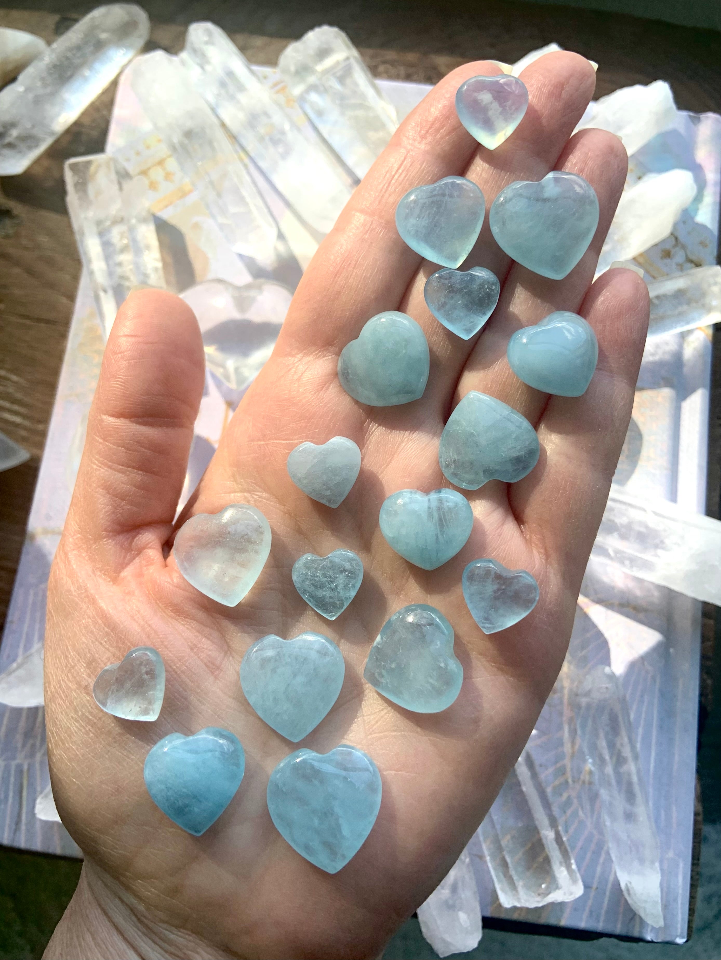 Aquamarine hearts displayed on a hand above a tabletop with quartz points — healing crystals for peace, emotional balance + open communication.

