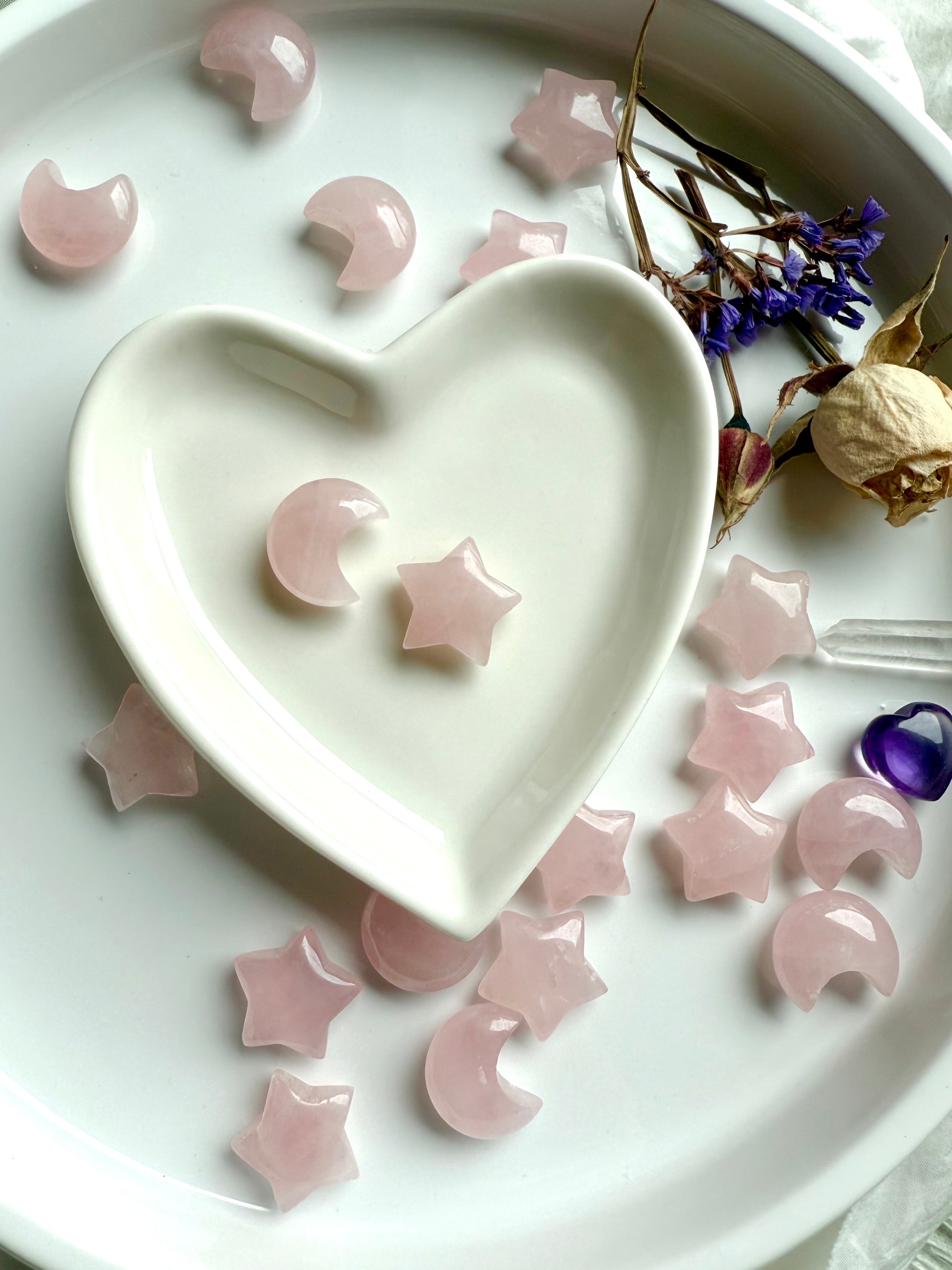 Mini Rose Quartz moon and star in a white heart bowl, surrounded by more moons and stars. Heart-centered carvings for peace, love & self-care rituals.