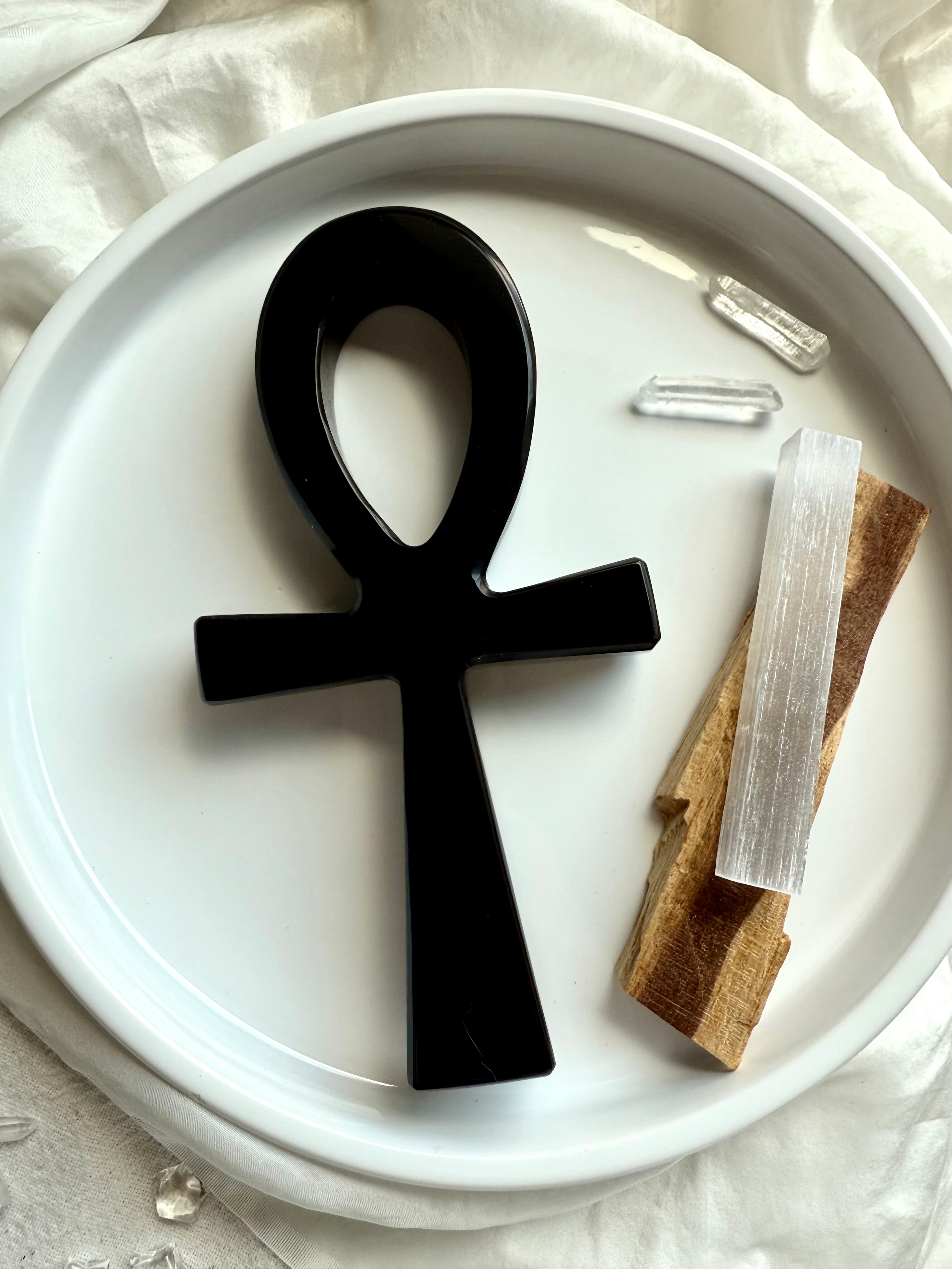 Flatlay of a Black ankh symbol on a white plate with crystals and palo santo sticks.