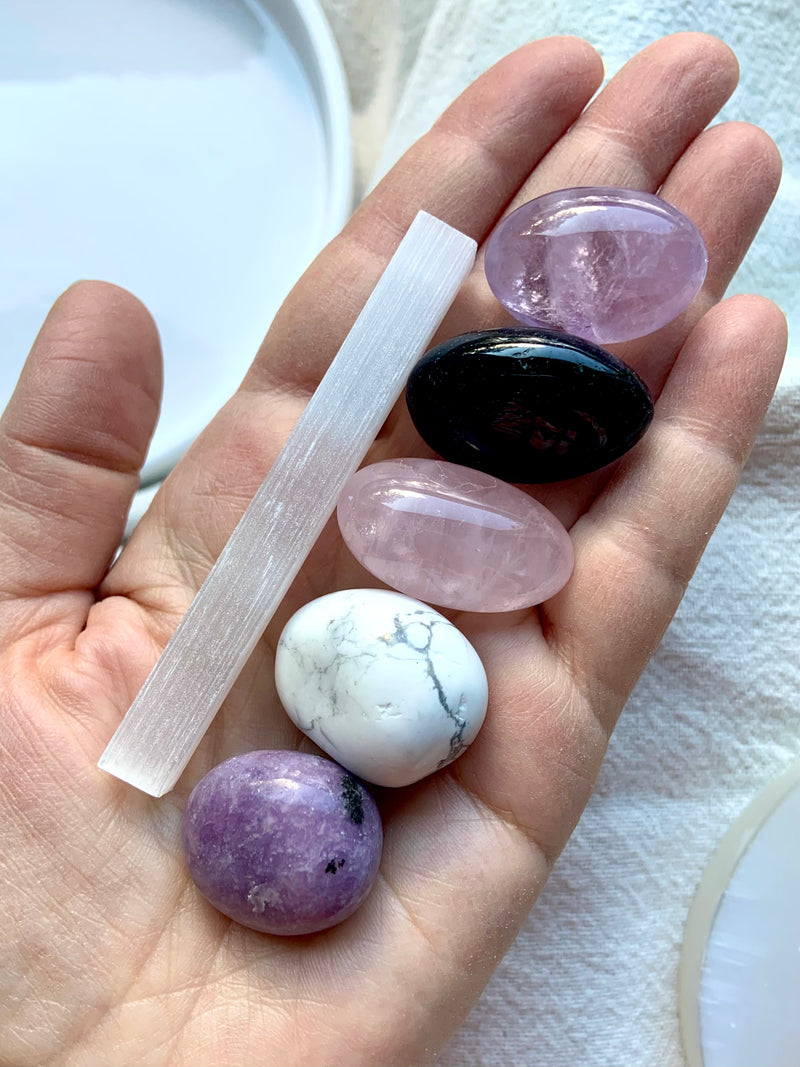 The Sweet Sleep Crystal Set to help you relax at bedtime, shown on my hand with a creamy white background. Set contains 6 crystals: lepidolite, amethyst, rose quartz, howlite, black tourmaline + selenite.