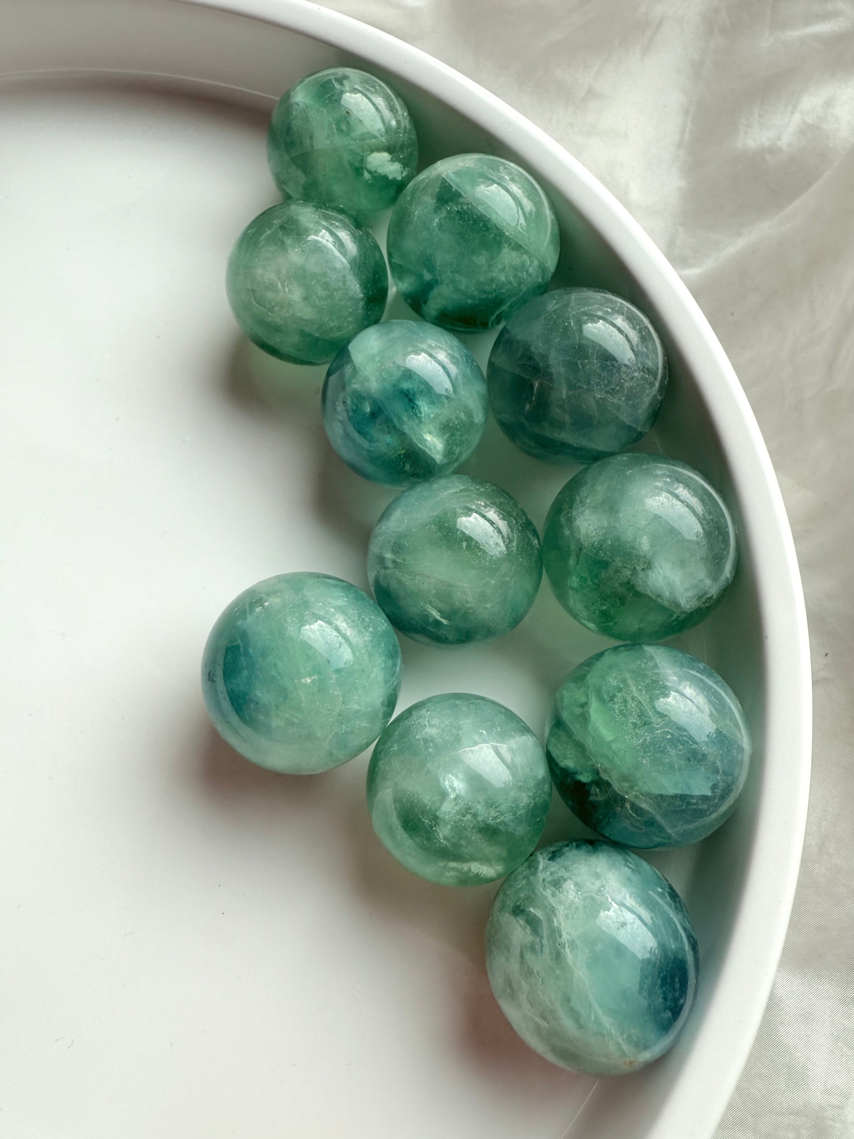 Teal Fluorite spheres with aqua green glow displayed on a white tray, mini crystal balls for clarity and throat chakra healing.