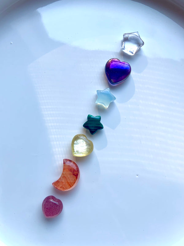 7 Chakra Crystal Set of Rainbow Gemstone Moons, Stars, and Heart Mini Carvings shown in a diagonal line on a white plate in natural light with shadows