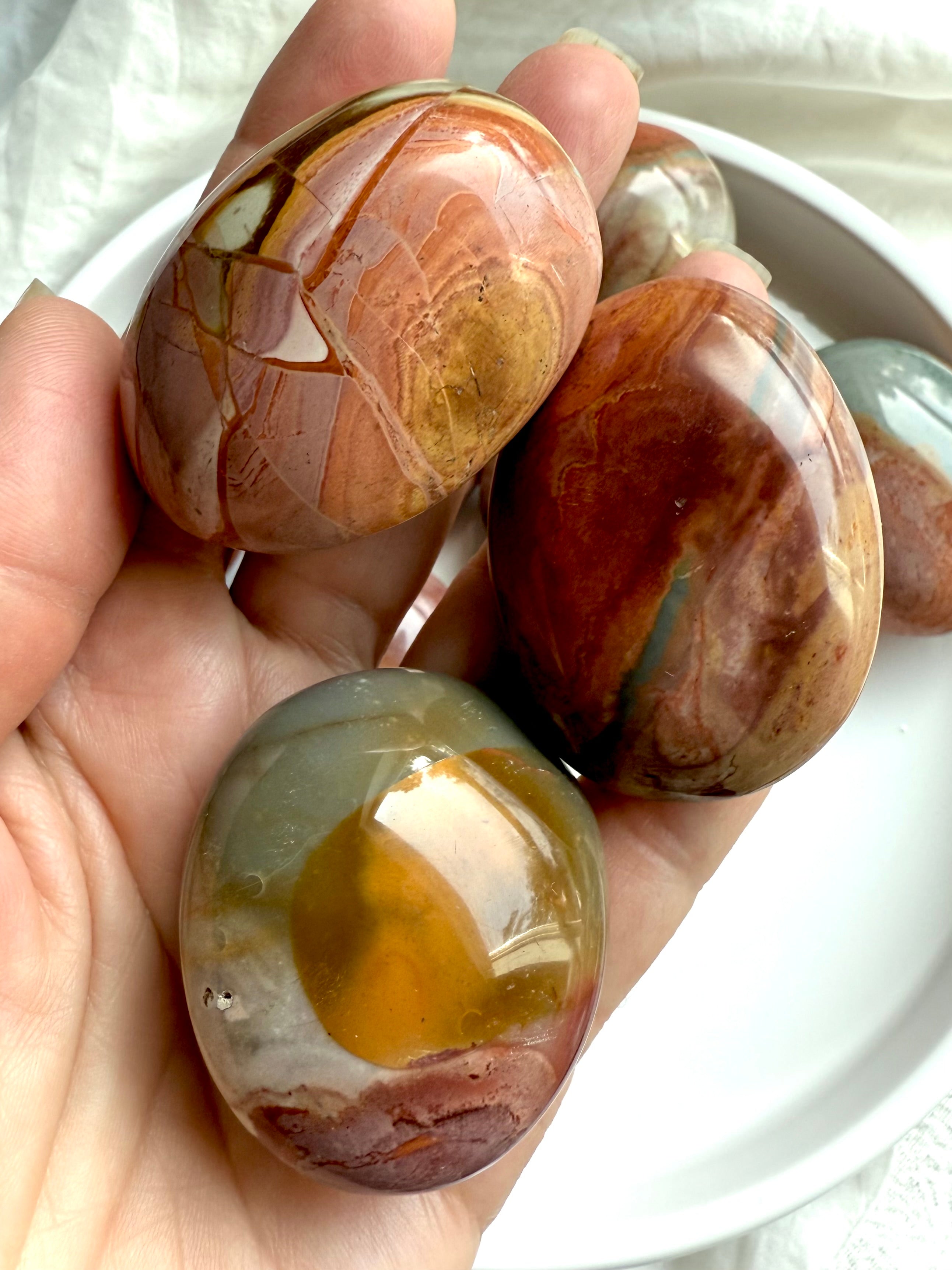 Polychrome Jasper palm stones in natural tones with swirling patterns shown on a hand. 