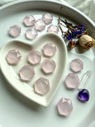 Angled view of rose quartz hexagon crystals in a white heart bowl with more scattered on a plate, styled with roses, clear quartz and amethyst heart