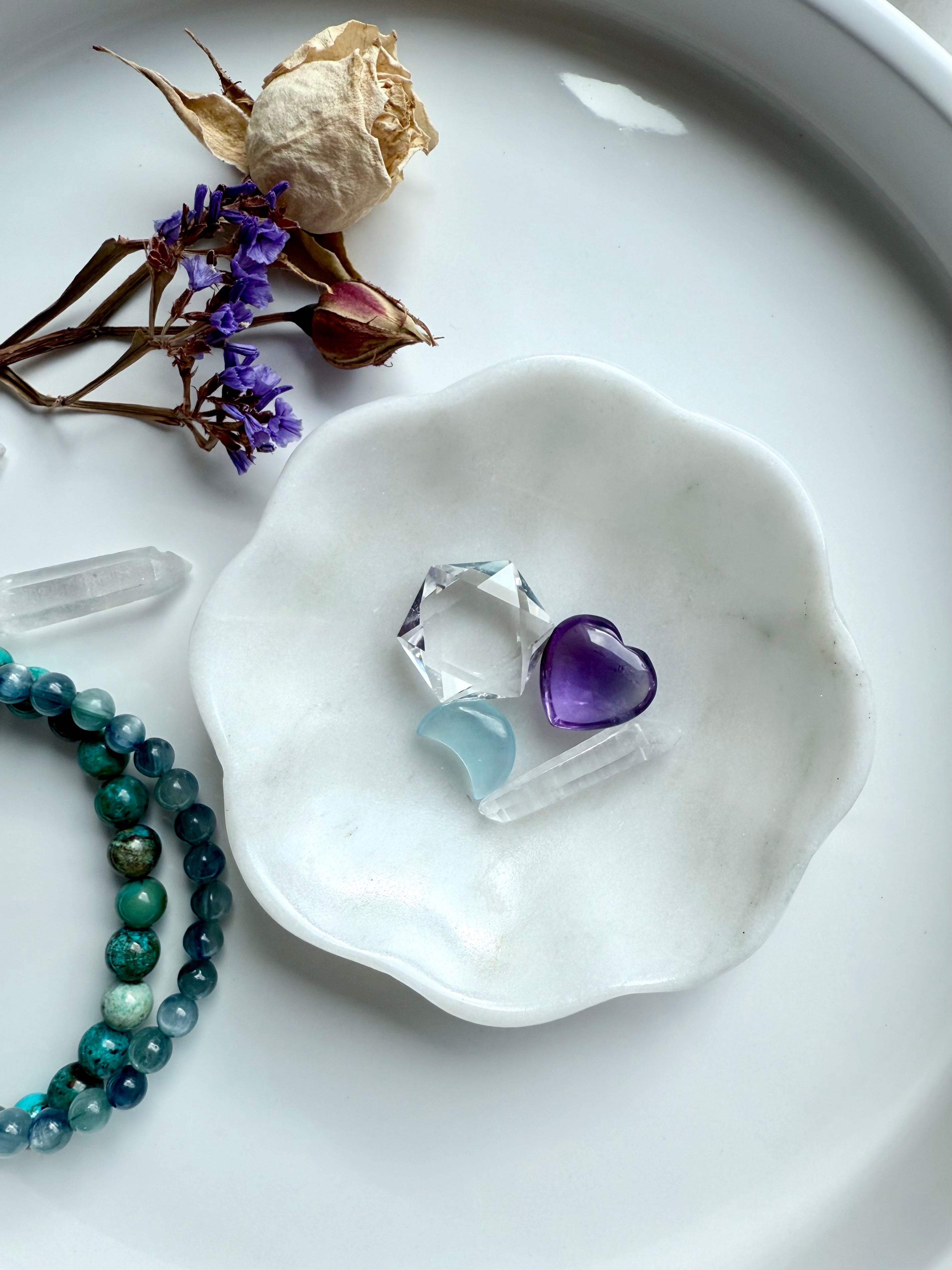 White marble daisy bowl with scalloped flower edges holding an amethyst heart carving, aquamarine moon, and a clear quartz hexagon
