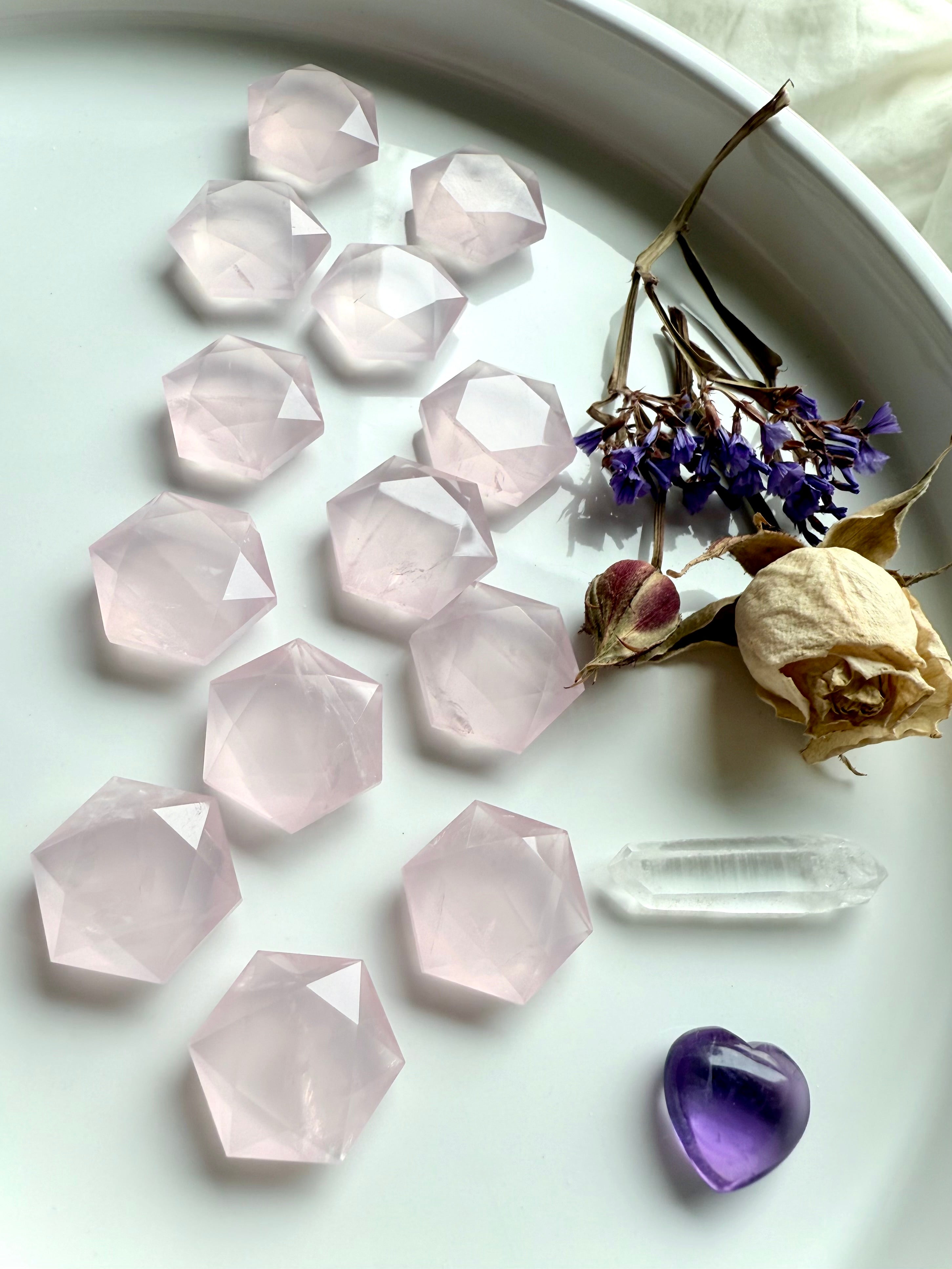 Flatlay of rose quartz hexagon crystals on a white plate styled with roses, clear quartz point and amethyst heart, sacred geometry carvings for grids