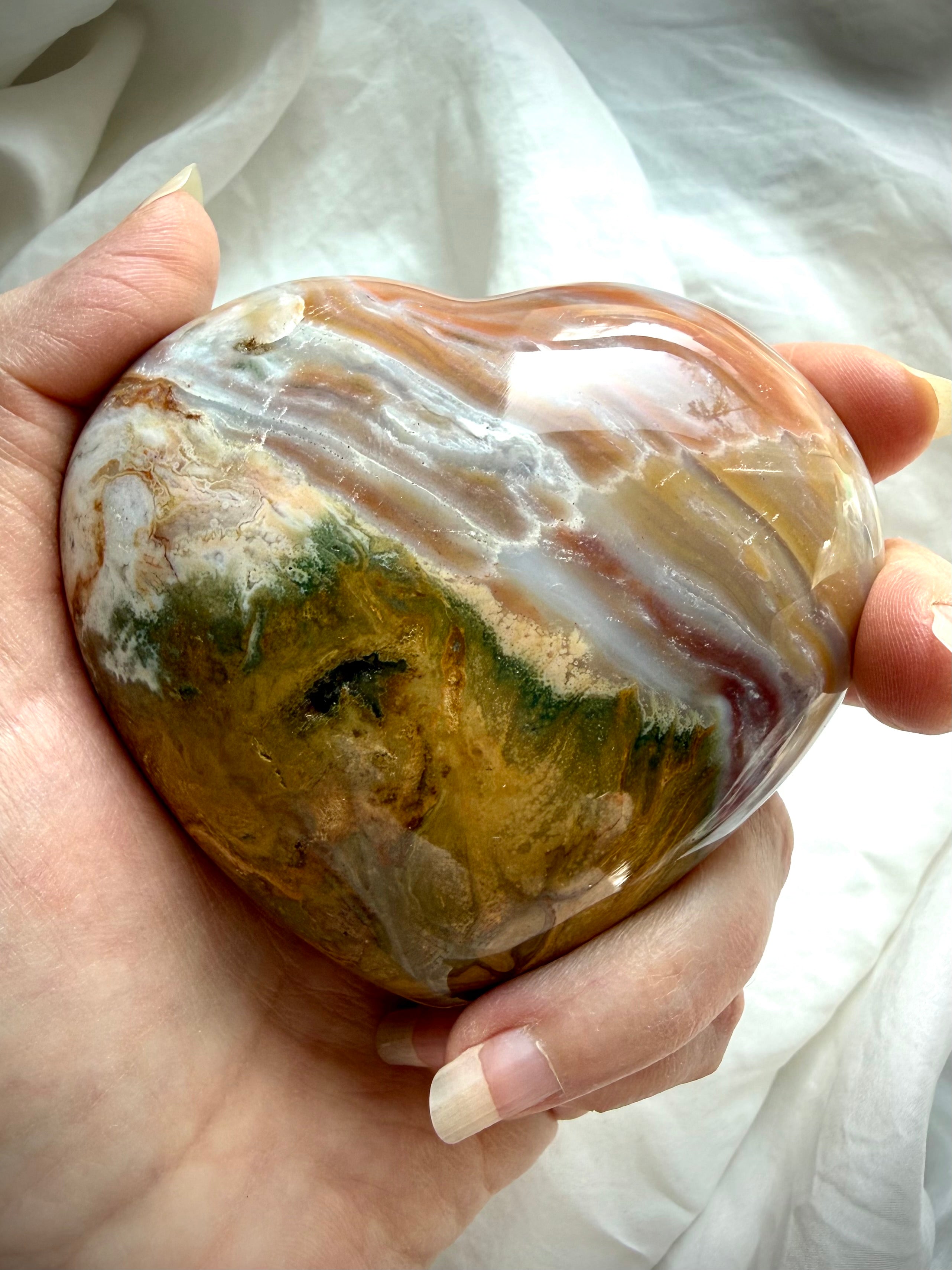 One of a kind Ocean Jasper heart crystal also known as sea jasper, with flowing agate banding in caramel, cream, moss green and soft grey tones, held in hand
