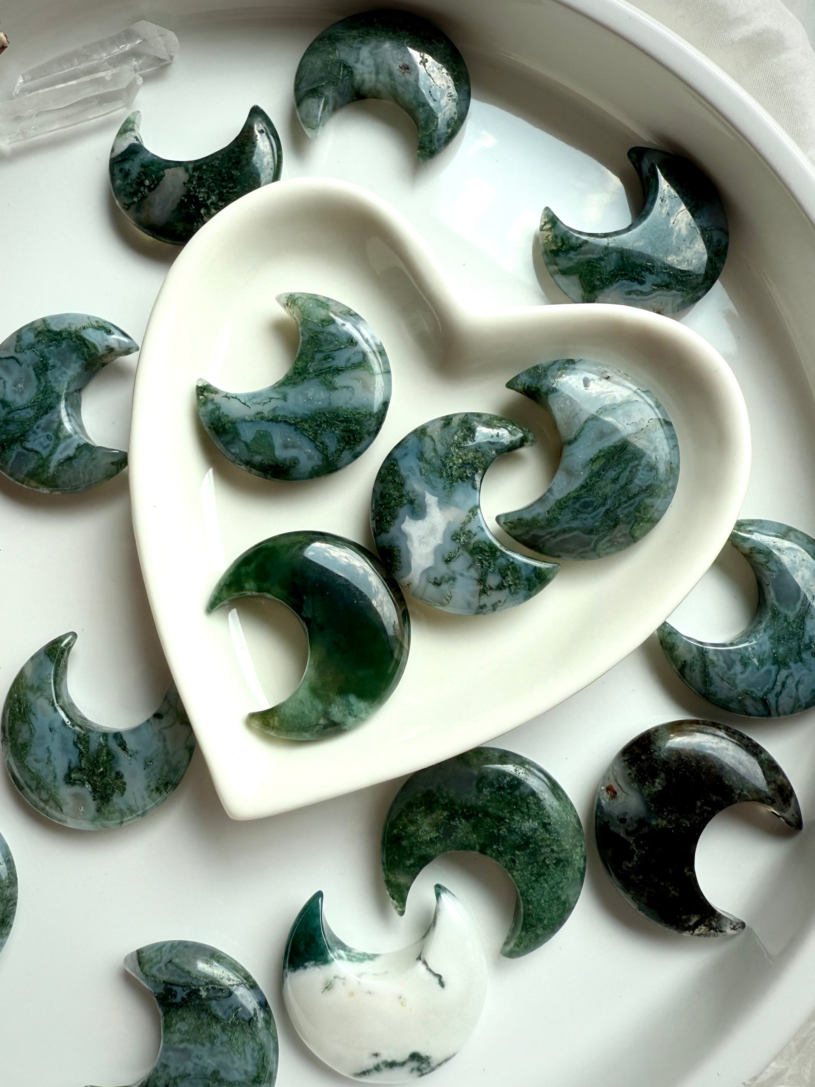 Moss Agate moon carvings in a white heart-shaped bowl with additional moons surrounding it, styled for gifting or altar display.