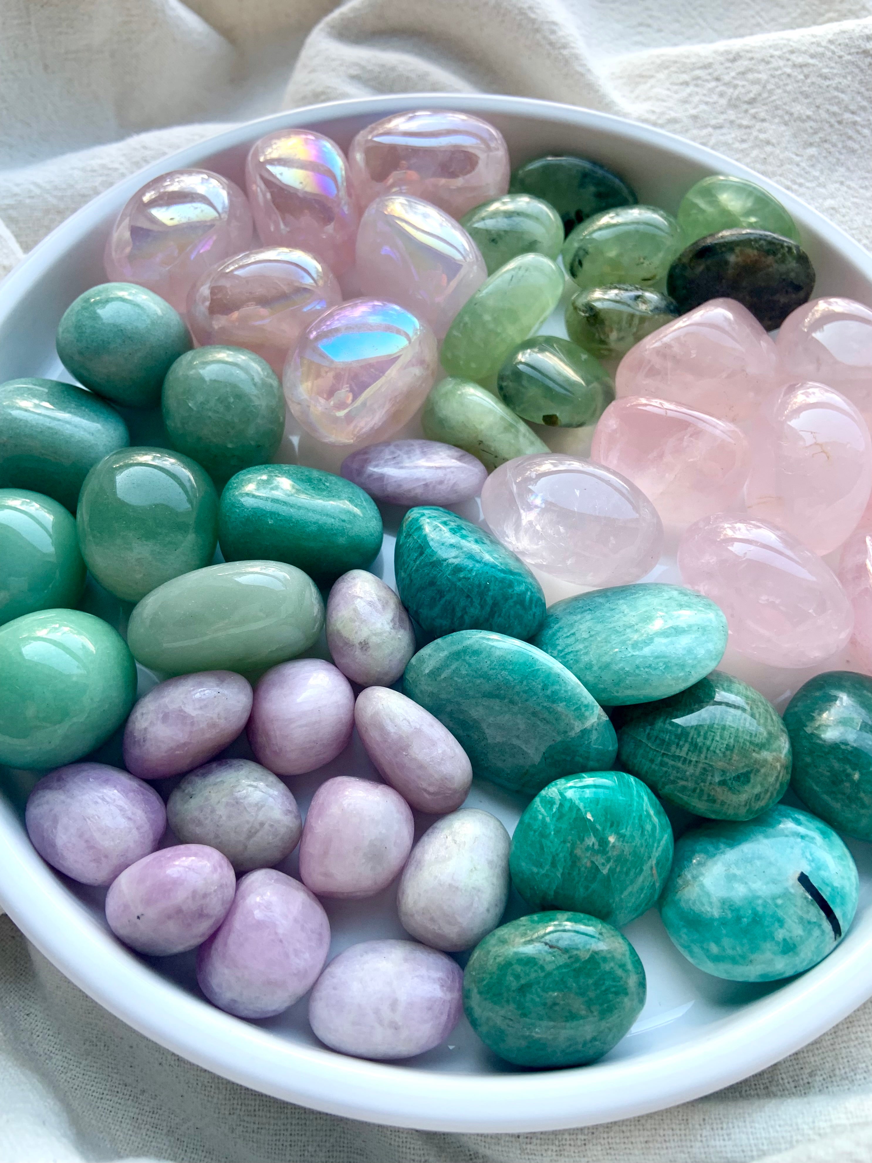 Assorted Heart Chakra healing crystals in pink and green arranged on a white plate — aura rose quartz, kunzite, amazonite, prehnite, rose quartz, and green aventurine.