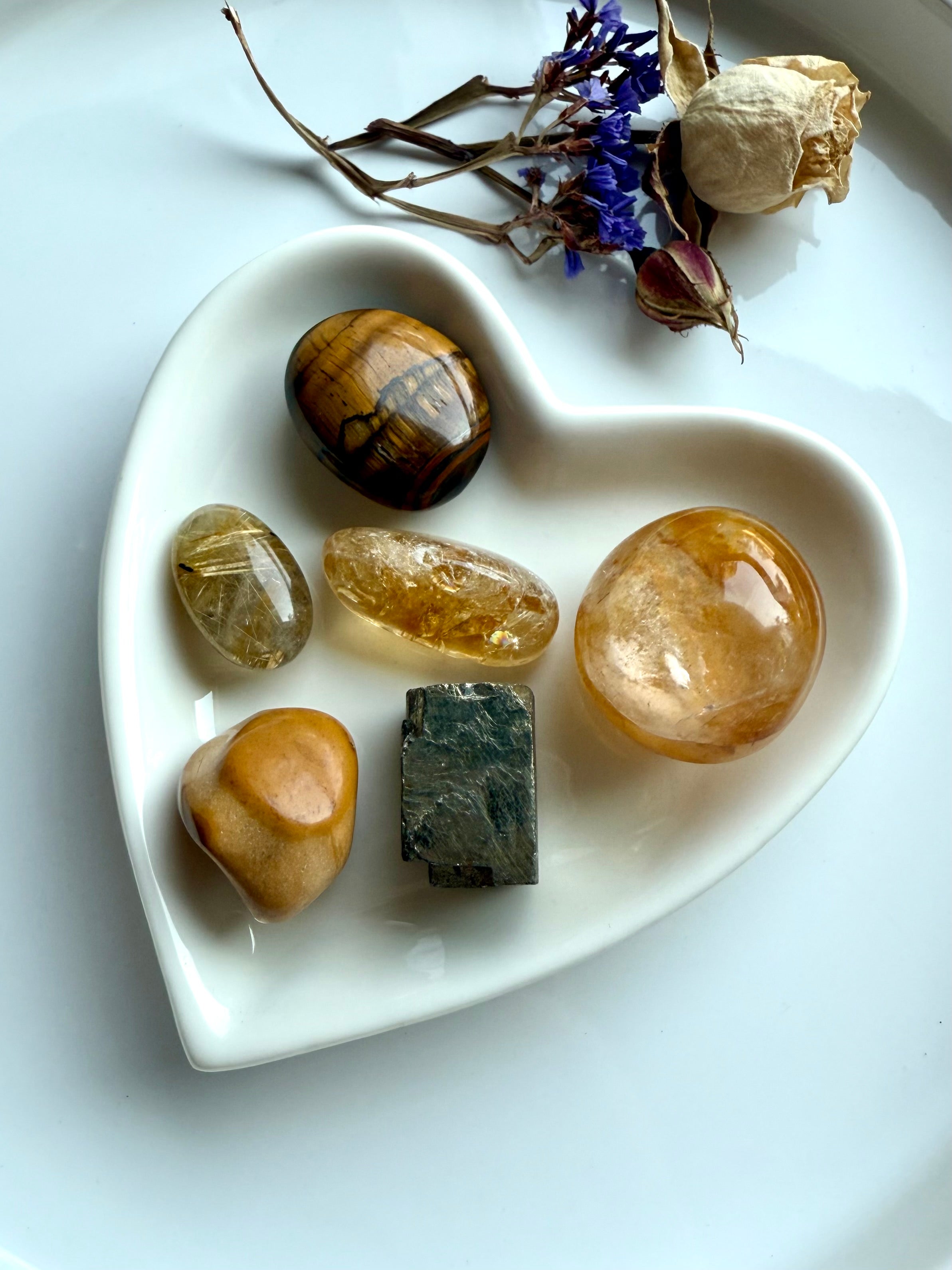 Solar Plexus Chakra crystal set arranged in a white heart-shaped bowl with dried flowers — golden healer, citrine, golden rutile, tiger’s eye, yellow mookaite, and raw pyrite