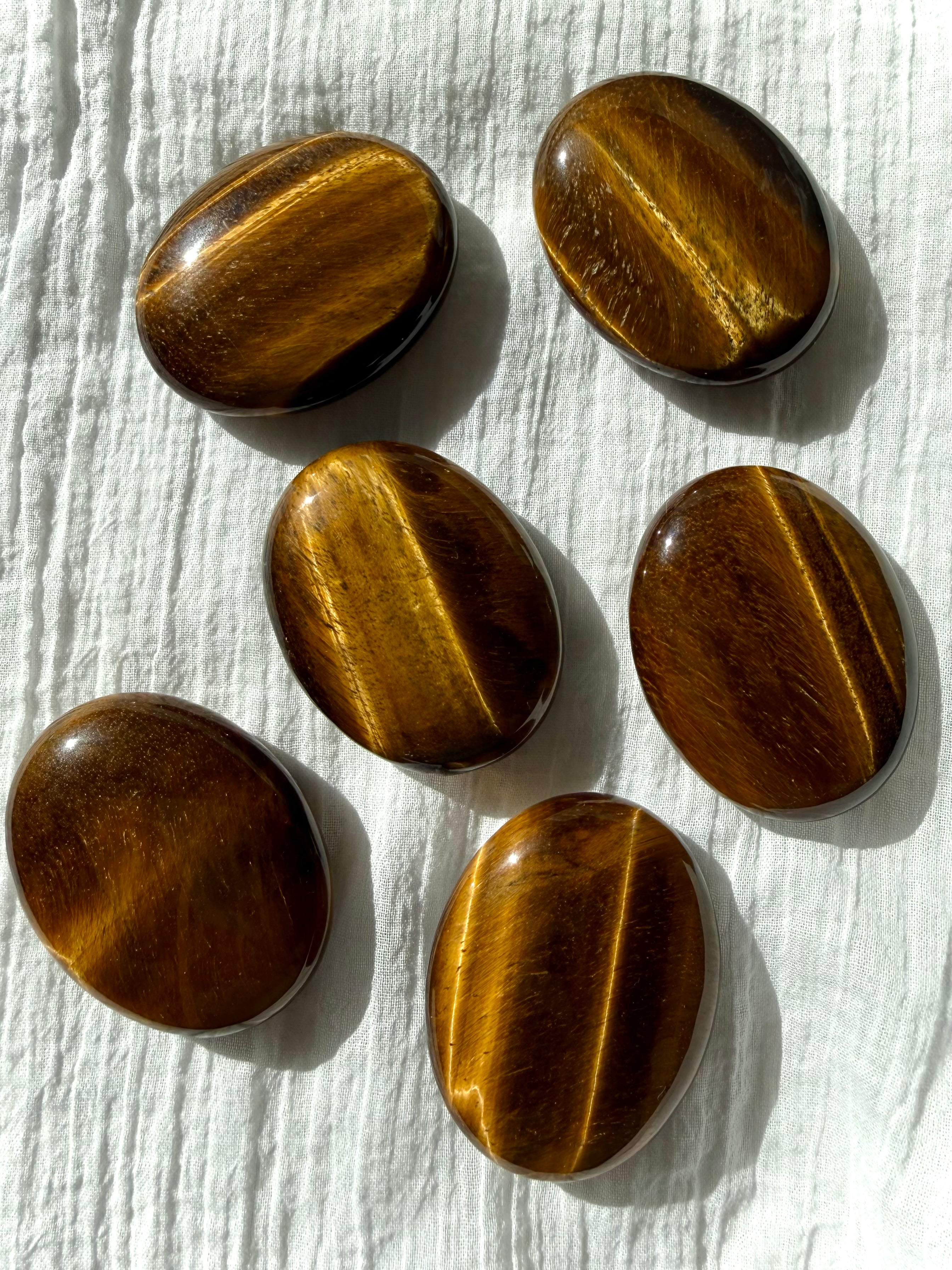 Tiger’s Eye palm stones arranged on white fabric showing beautiful golden flash and earthy brown tones