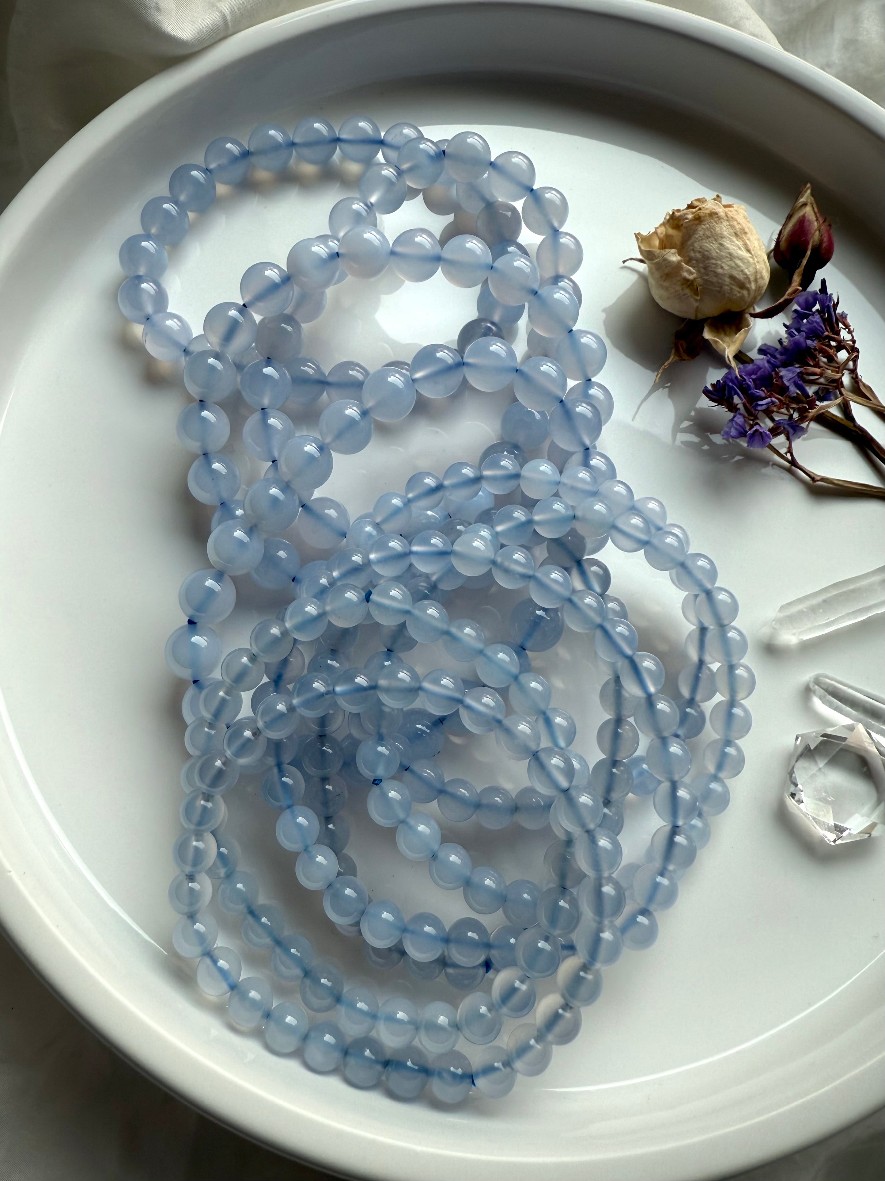Glossy Blue Chalcedony bracelet strands with dried roses, statice flowers, and clear quartz points on white dish