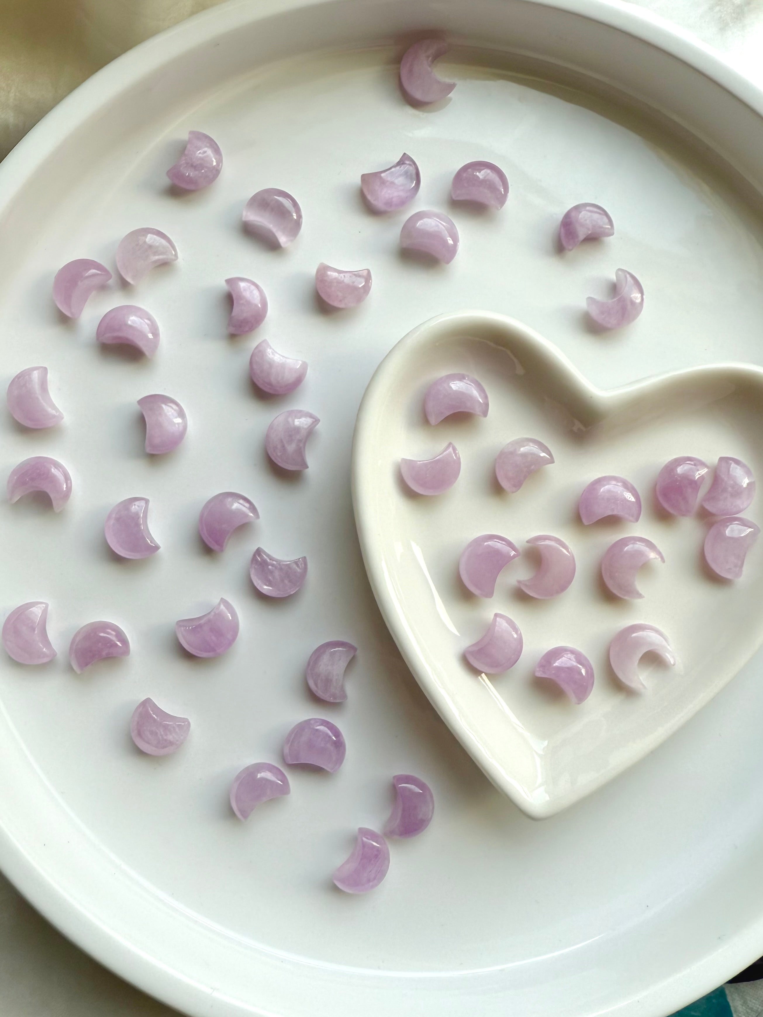 Pink Kunzite crescent moon carvings in a heart-shaped bowl with more moons scattered on a white plate — soothing crystals for heart chakra healing.