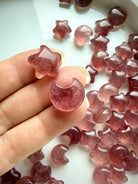 Mini Strawberry Quartz crescent moon held between fingers, showing translucent rosy pink color and natural red mineral inclusions