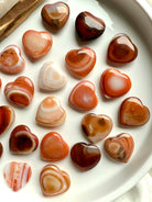 Mini Carnelian heart crystals arranged on white tray, showcasing natural banding and warm orange tones for Sacral Chakra healing